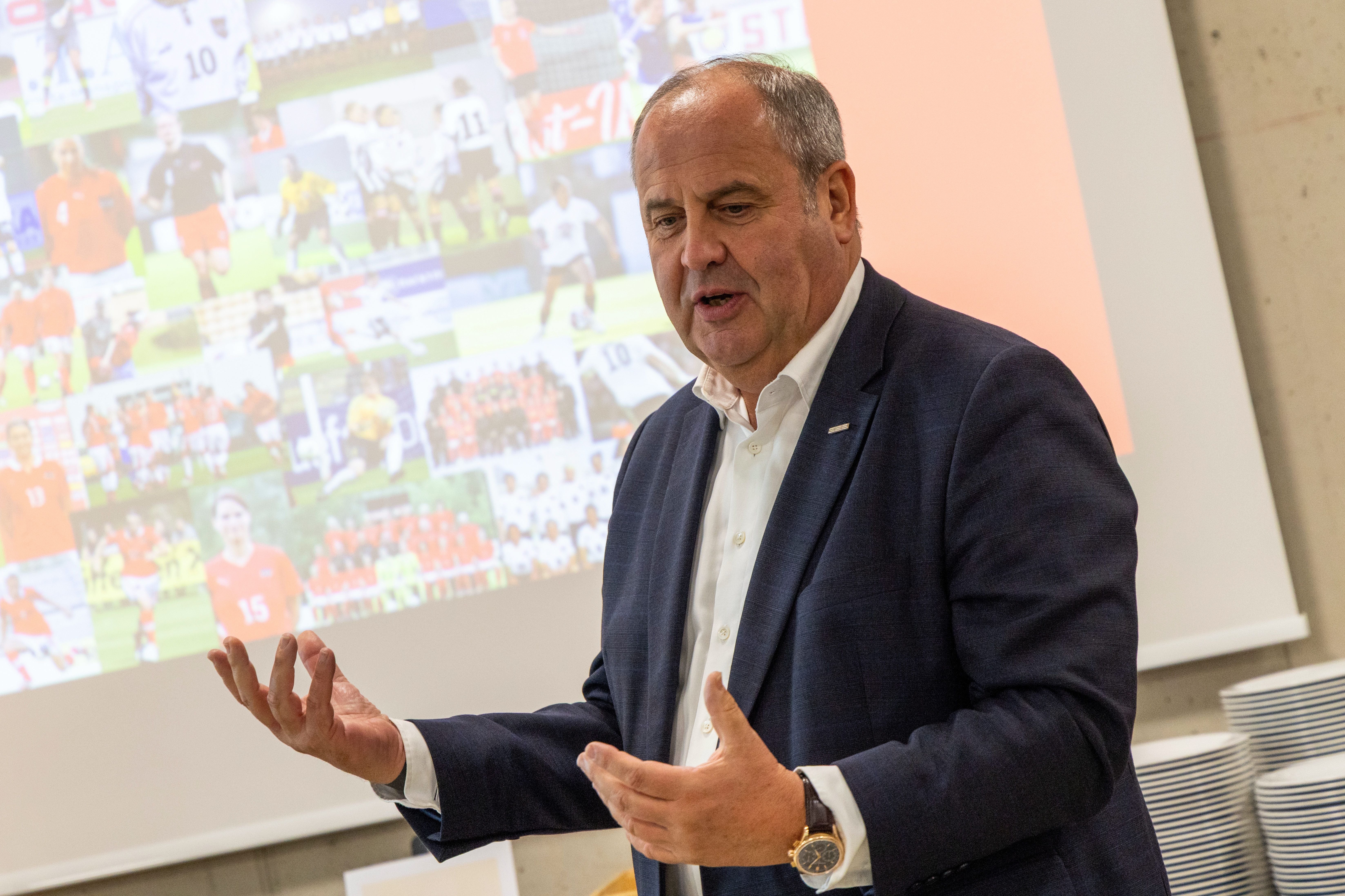 Ein Mann im Anzug spricht vor einer Leinwand mit Fußballfotos im Hintergrund.
