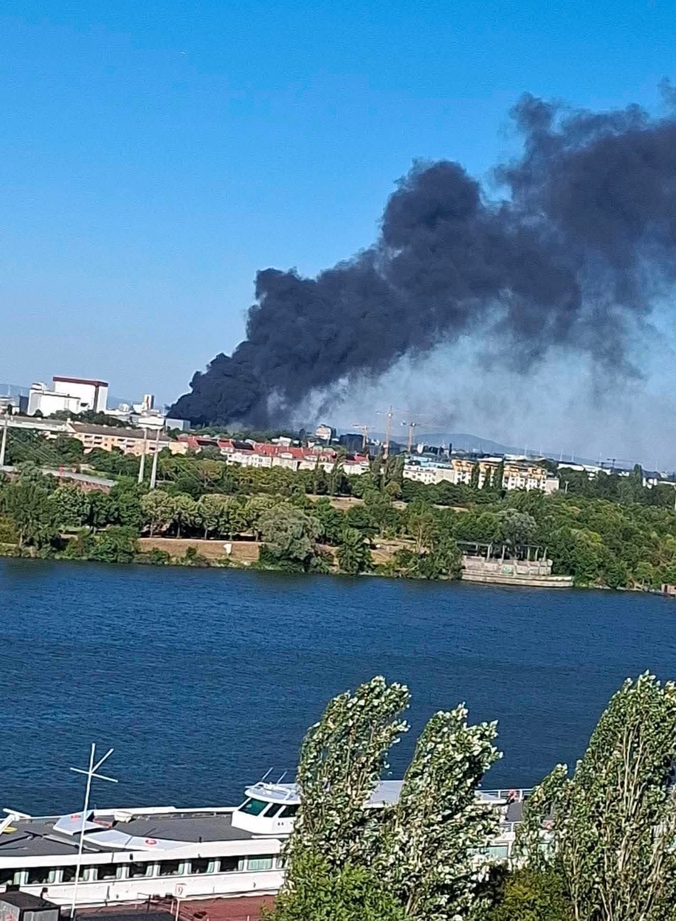Dichte schwarze Rauchwolke steigt über einem Industriegebiet bei einem Fluss auf.