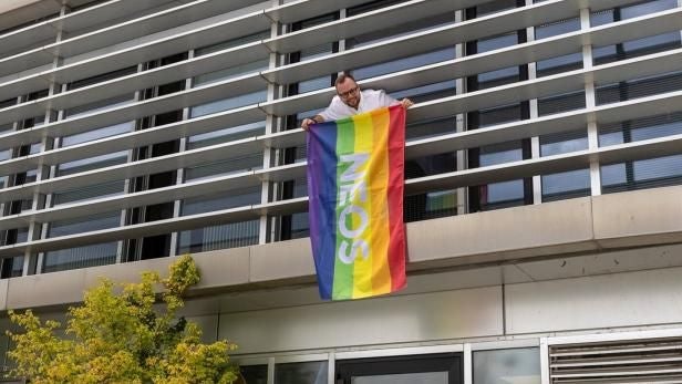 Ein Mann hängt eine Regenbogenflagge mit dem Schriftzug „NEOS“ aus einem Bürofenster.