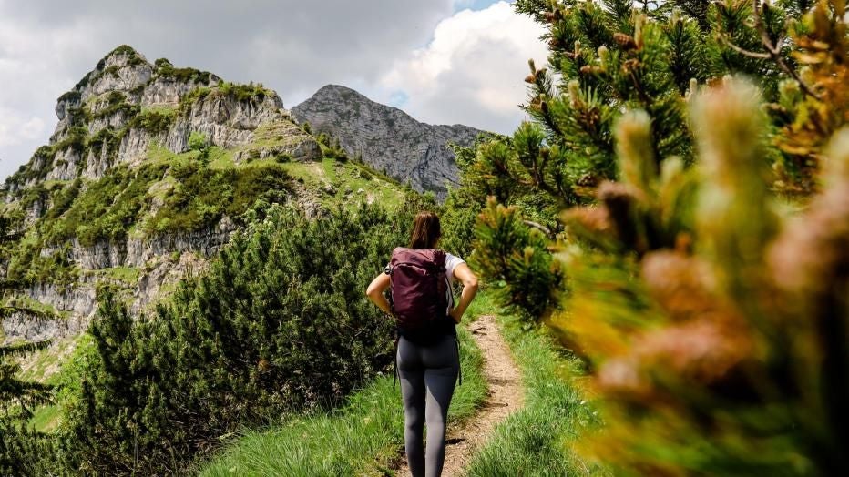Frau wandert auf einem schmalen Bergpfad zwischen grünen Büschen und Felsen.