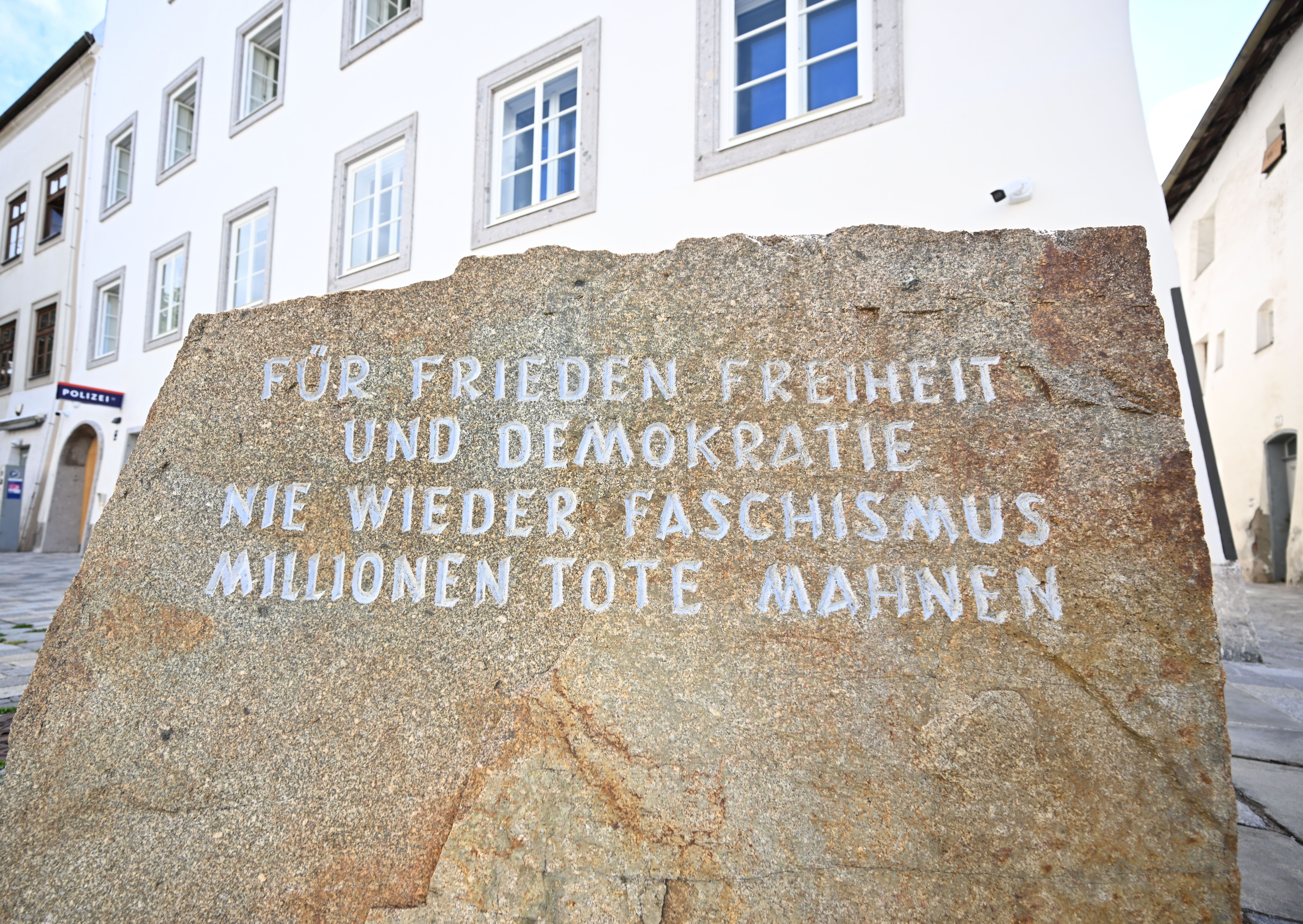 Gedenkstein mit Inschrift gegen Faschismus vor einem weißem Gebäude mit vielen Fenstern.