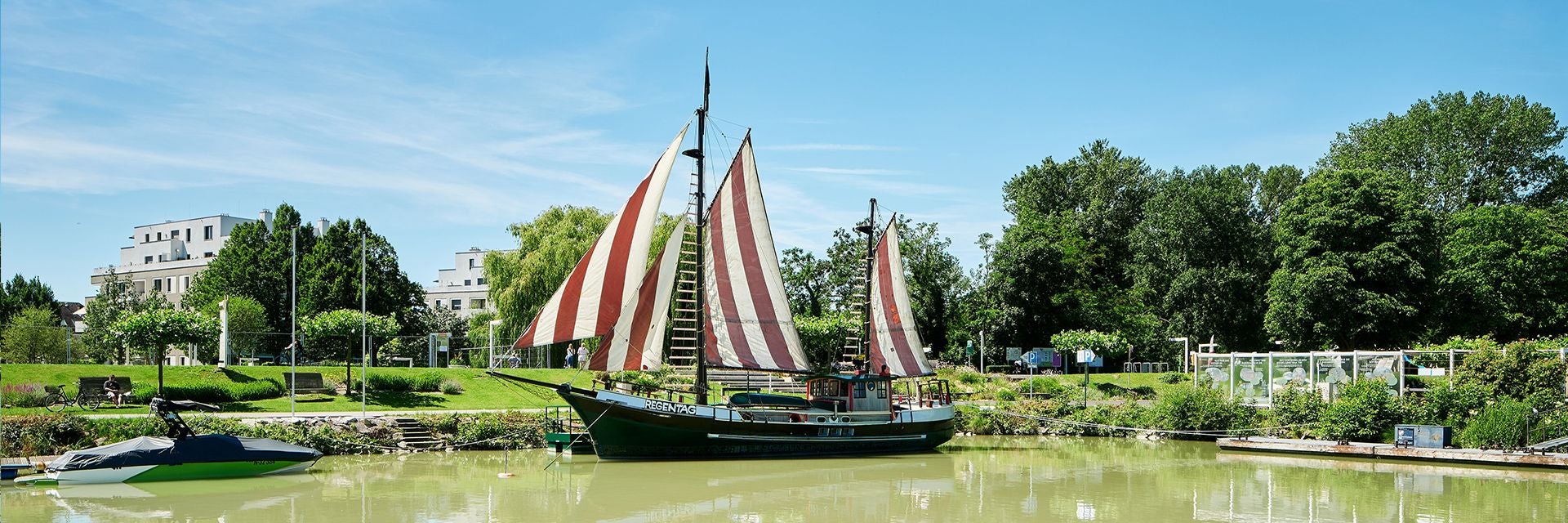 Segelschiff „Regentag“ mit rot-weißen Segeln auf einem ruhigen Gewässer vor Bäumen und Gebäuden.