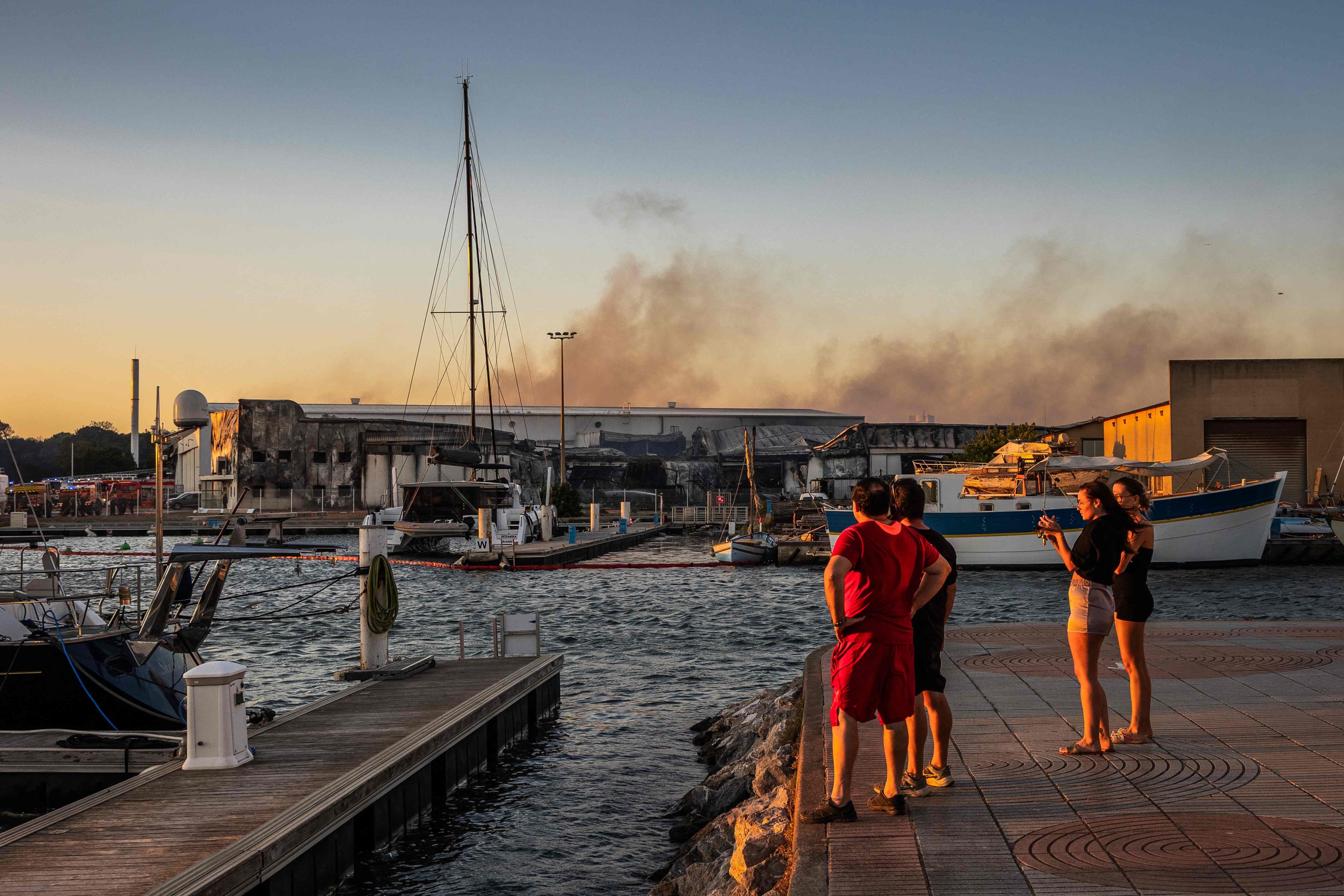 Mehrere Menschen beobachten am Hafen Rauch, der aus brennenden Gebäuden im Hintergrund aufsteigt.