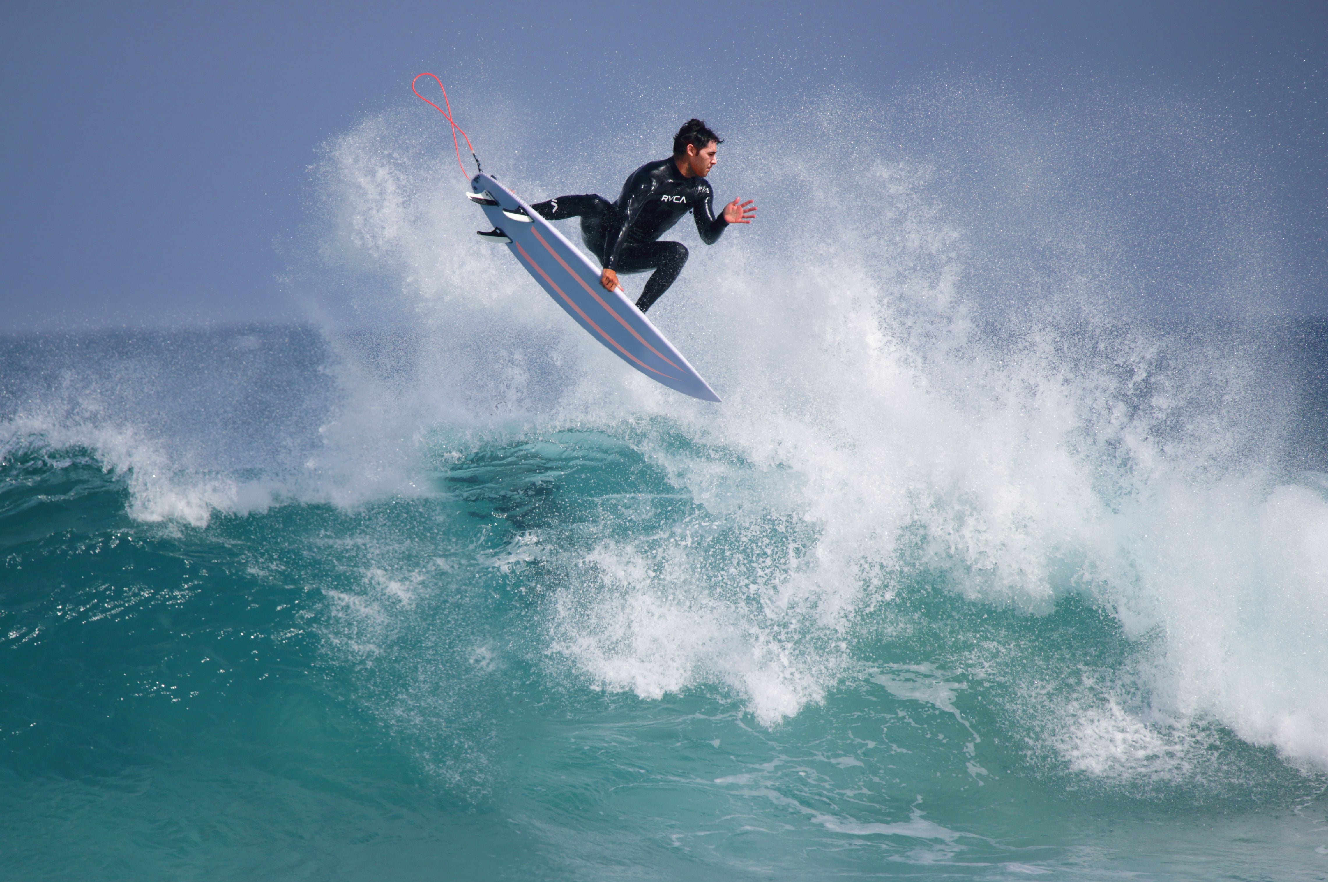 Ein Surfer springt mit seinem Brett über eine Welle im türkisfarbenen Meer.