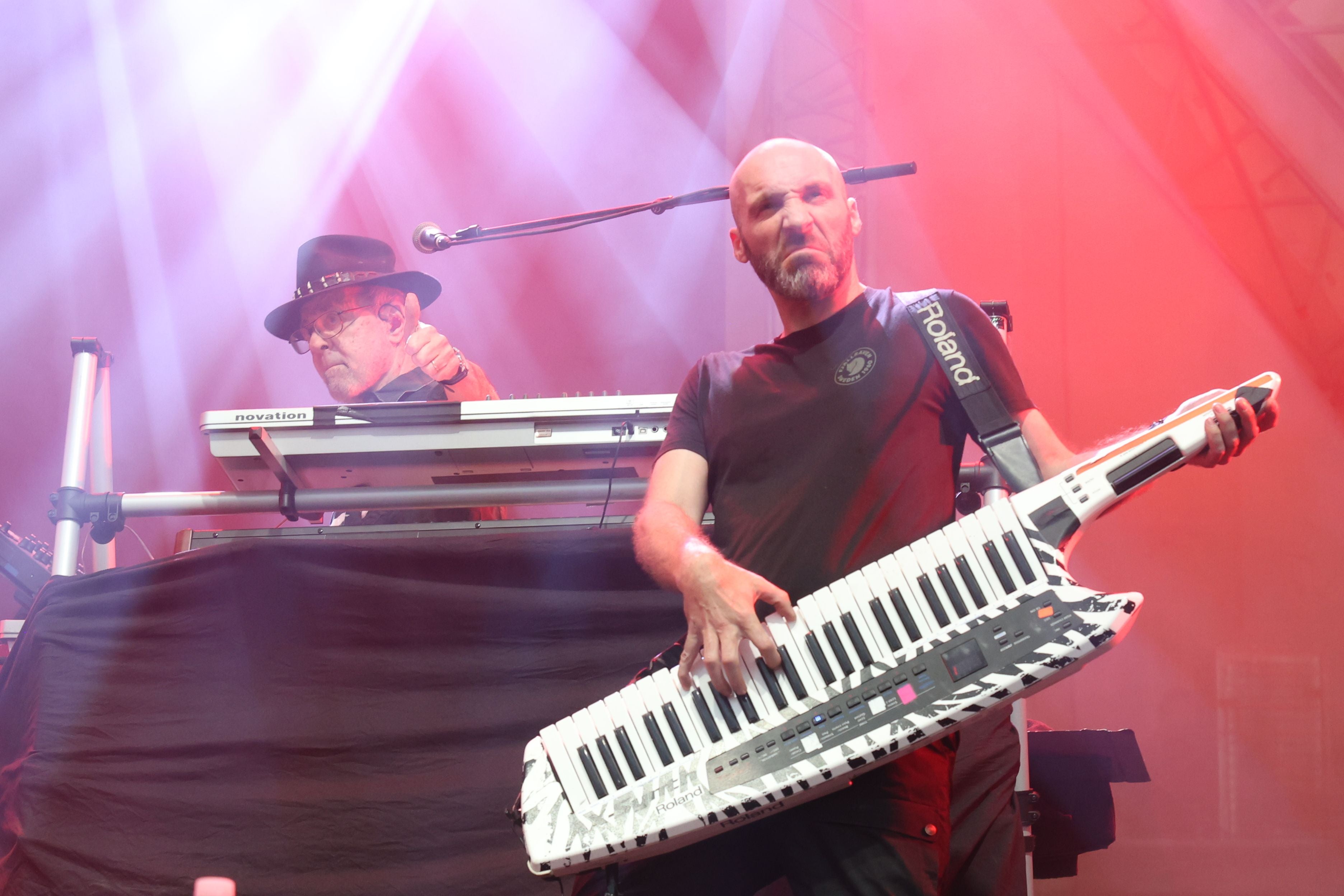 Zwei Musiker spielen Keyboards auf einer Bühne mit roter Beleuchtung und Nebel.