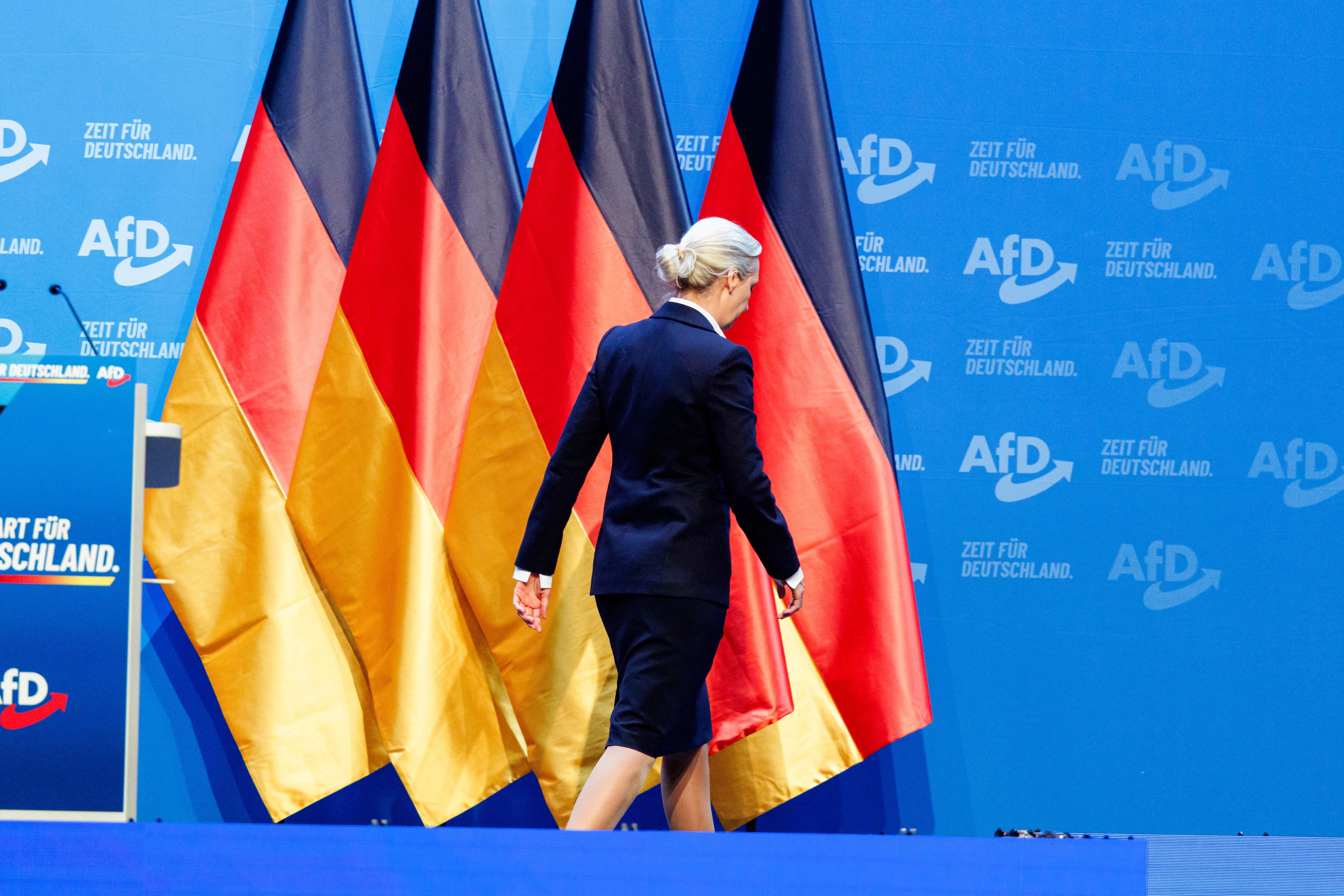 Frau im Anzug verlässt Bühne vor Deutschlandflaggen und AfD-Logos auf blauem Hintergrund.