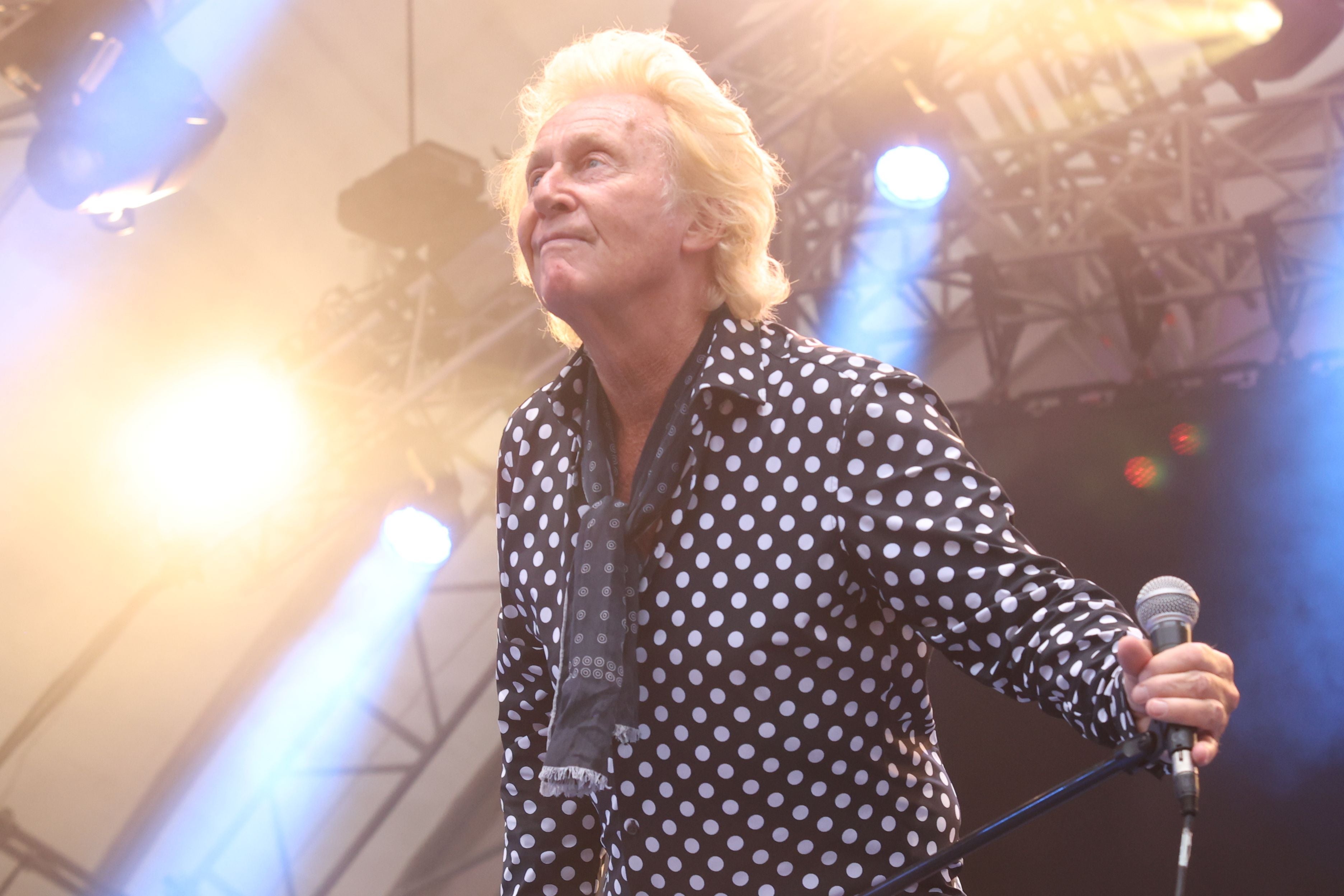 Ein älterer Mann mit blondem Haar singt auf einer Bühne mit Mikrofon und Lichtstrahlern.