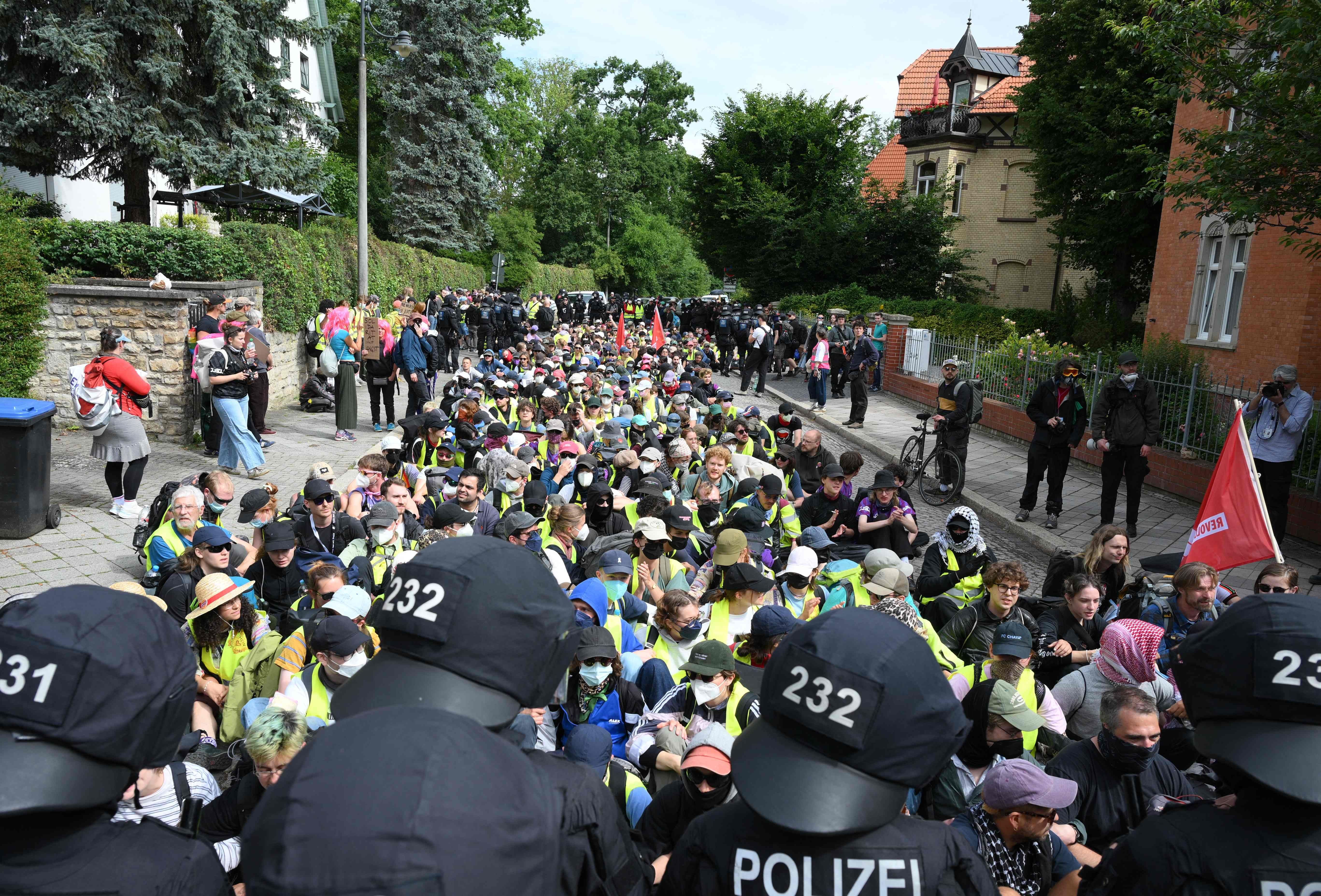 Viele Demonstrierende blockieren sitzend eine Straße, umgeben von Polizei in Ausrüstung.