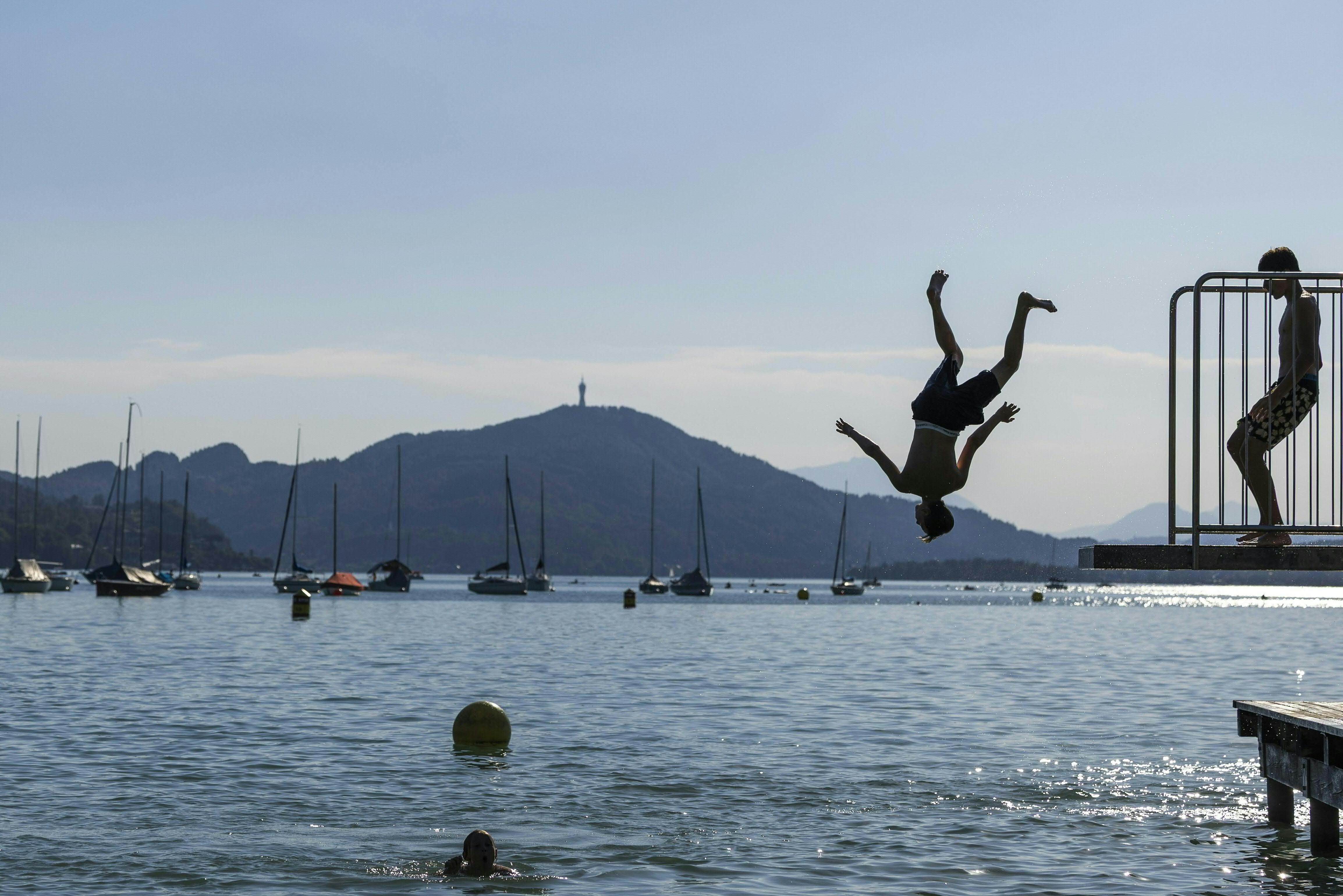 Junge springt kopfüber ins Wasser am Wörthersee, Boote und Berge im Hintergrund.
