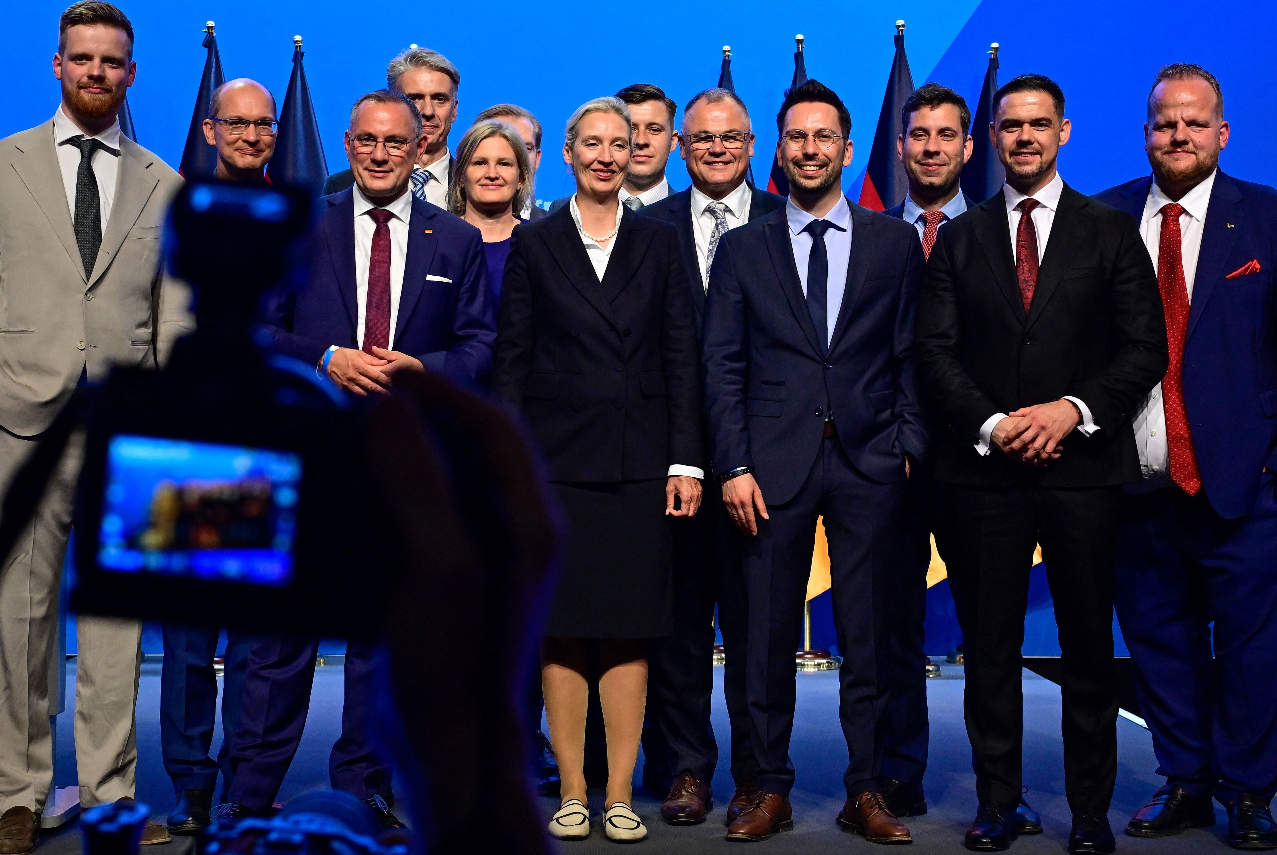 Eine Gruppe Menschen in Anzügen posiert auf einer Bühne, vorne ist eine Kamera sichtbar.