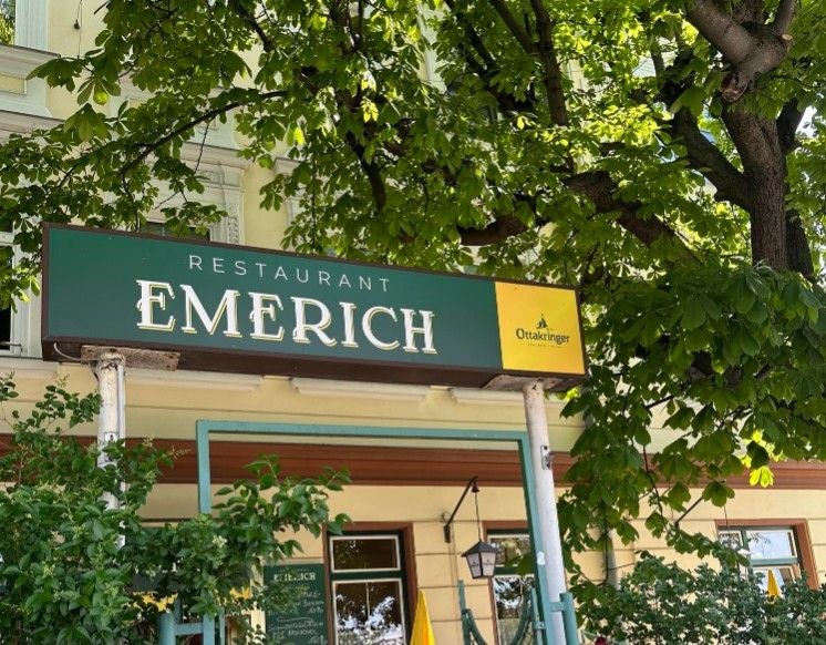 Grünes Schild mit der Aufschrift "Restaurant Emerich" vor einem Gebäude mit Bäumen.