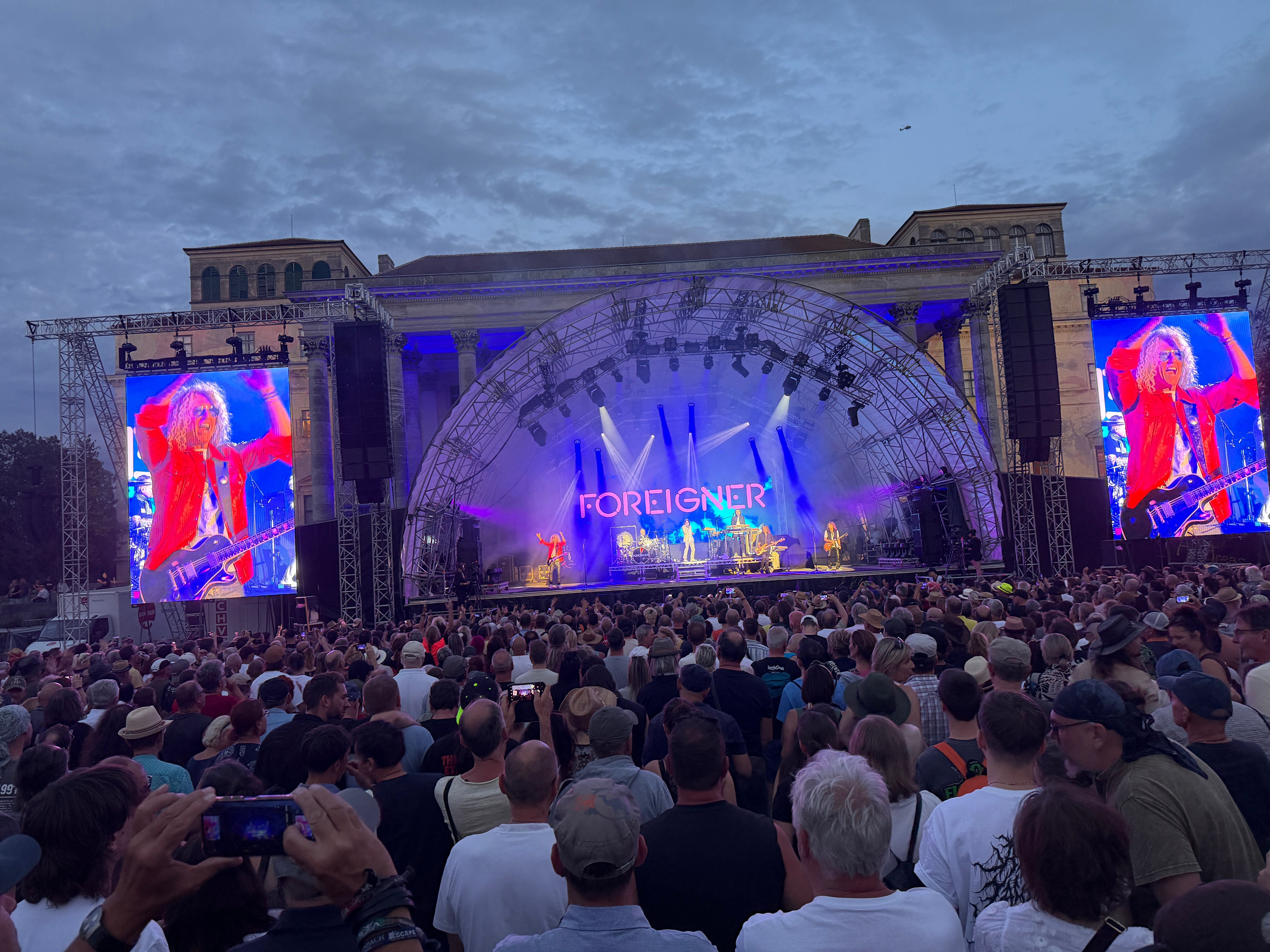 Große Menschenmenge vor einer Konzertbühne der Band Foreigner im Freien bei Abenddämmerung.