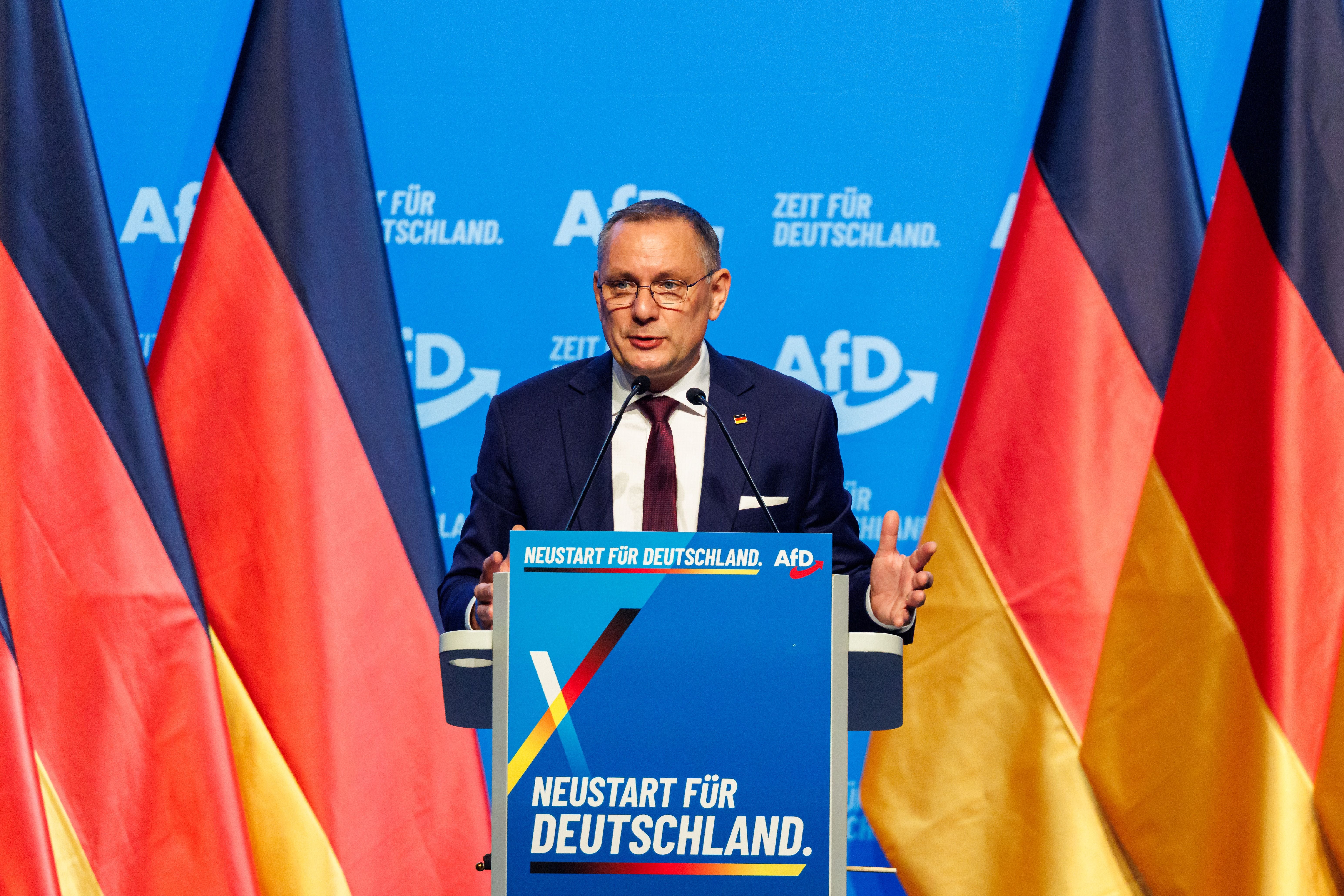 Ein Mann spricht am Rednerpult auf einem AfD-Parteitag, umgeben von deutschen Flaggen.