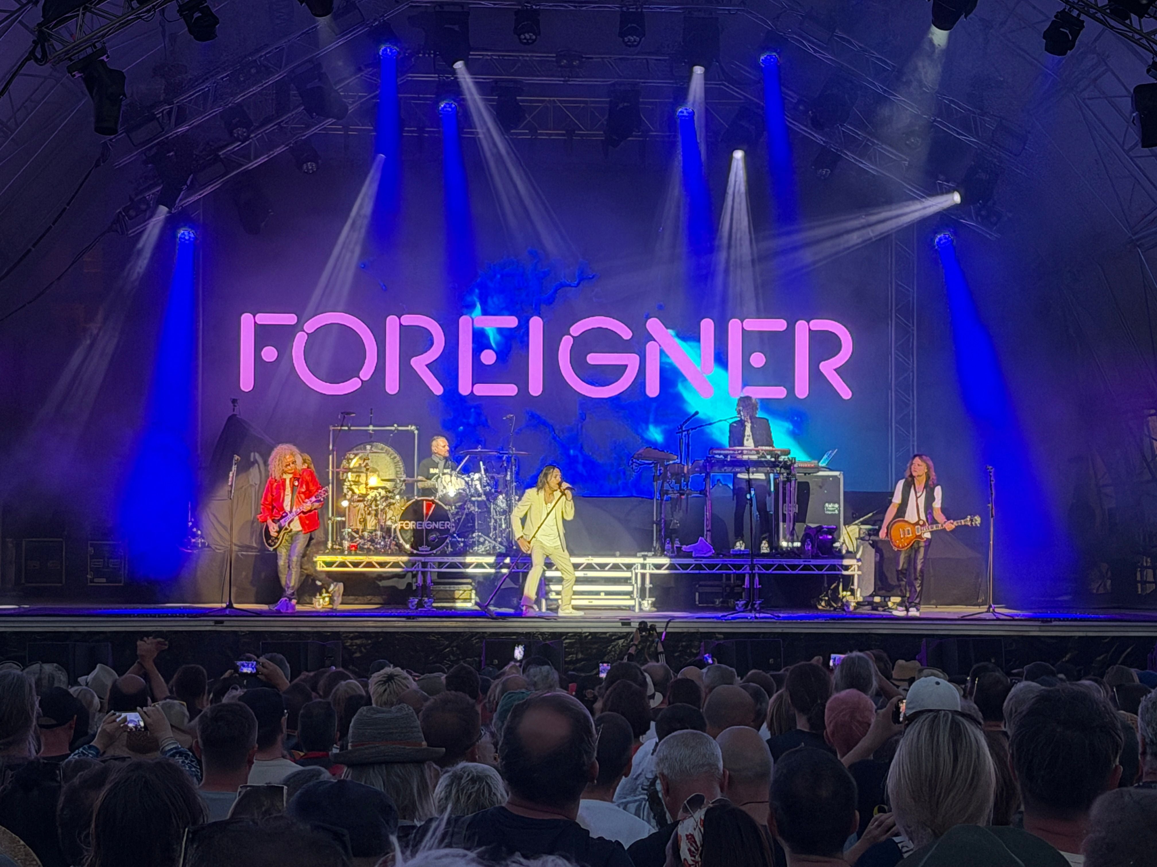 Die Band Foreigner tritt auf einer beleuchteten Bühne vor großem Publikum auf.
