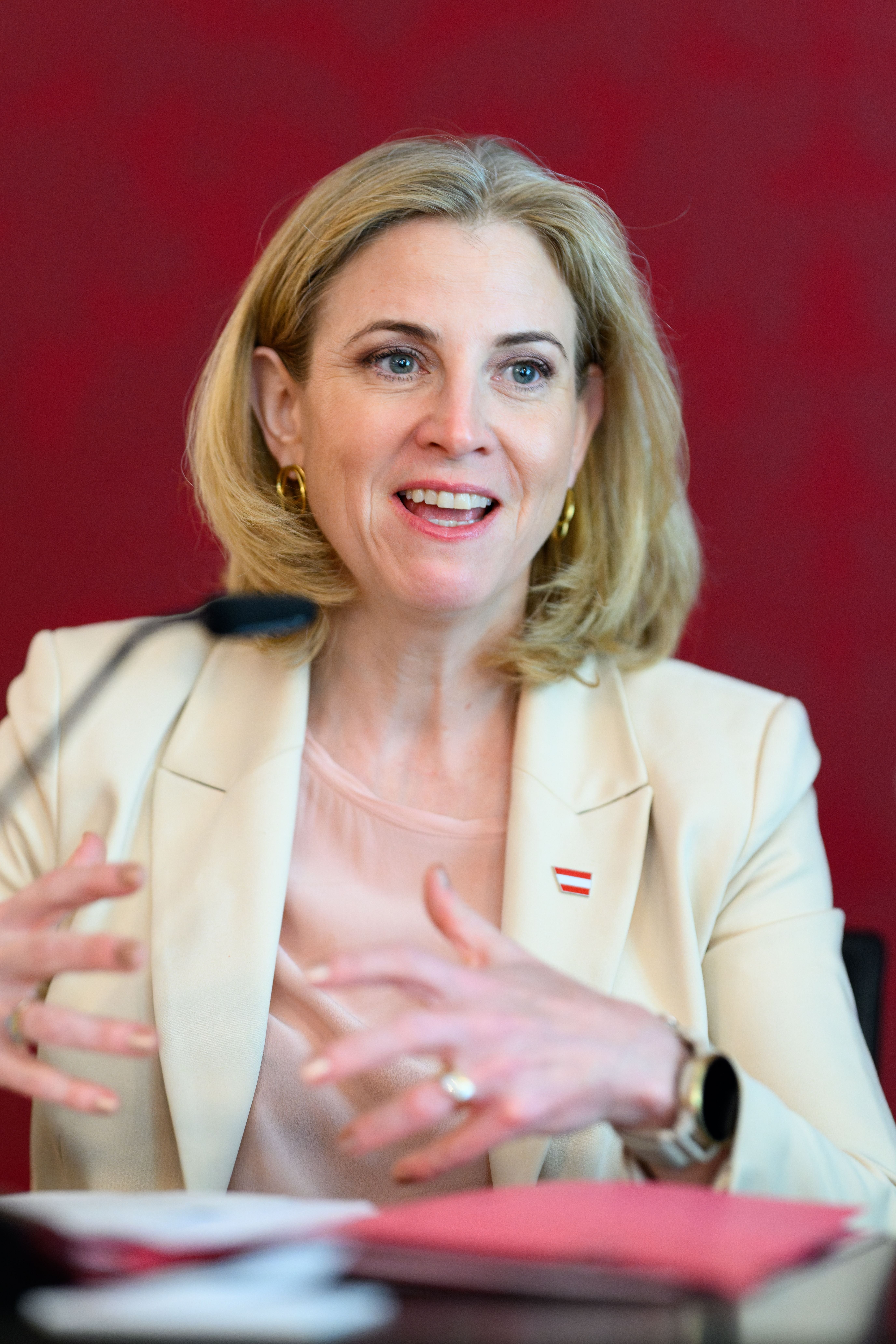 Eine Frau in hellem Blazer spricht gestikulierend vor rotem Hintergrund.