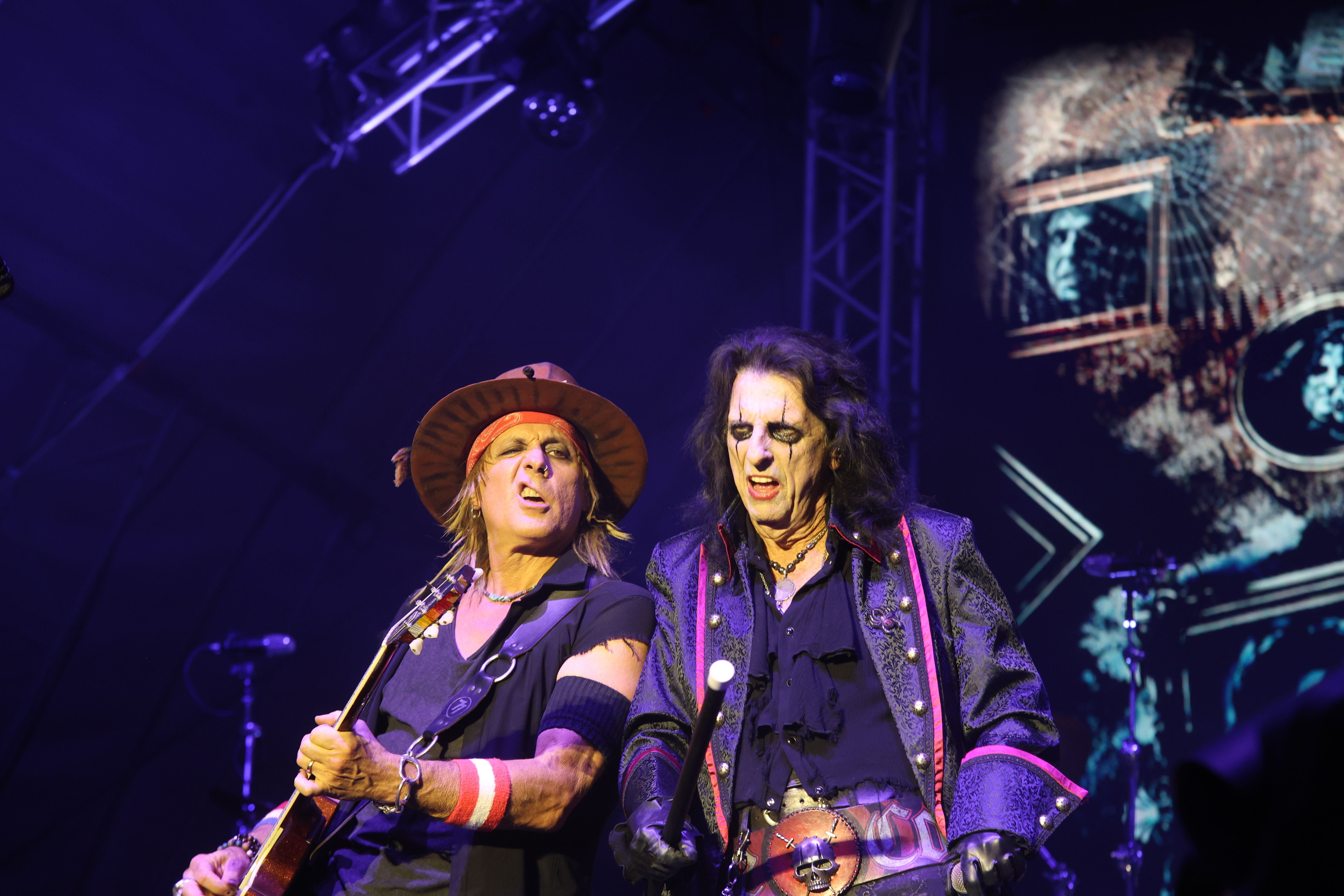 Zwei Rockmusiker mit Gitarren auf einer Bühne bei einem Konzert mit Lichteffekten.