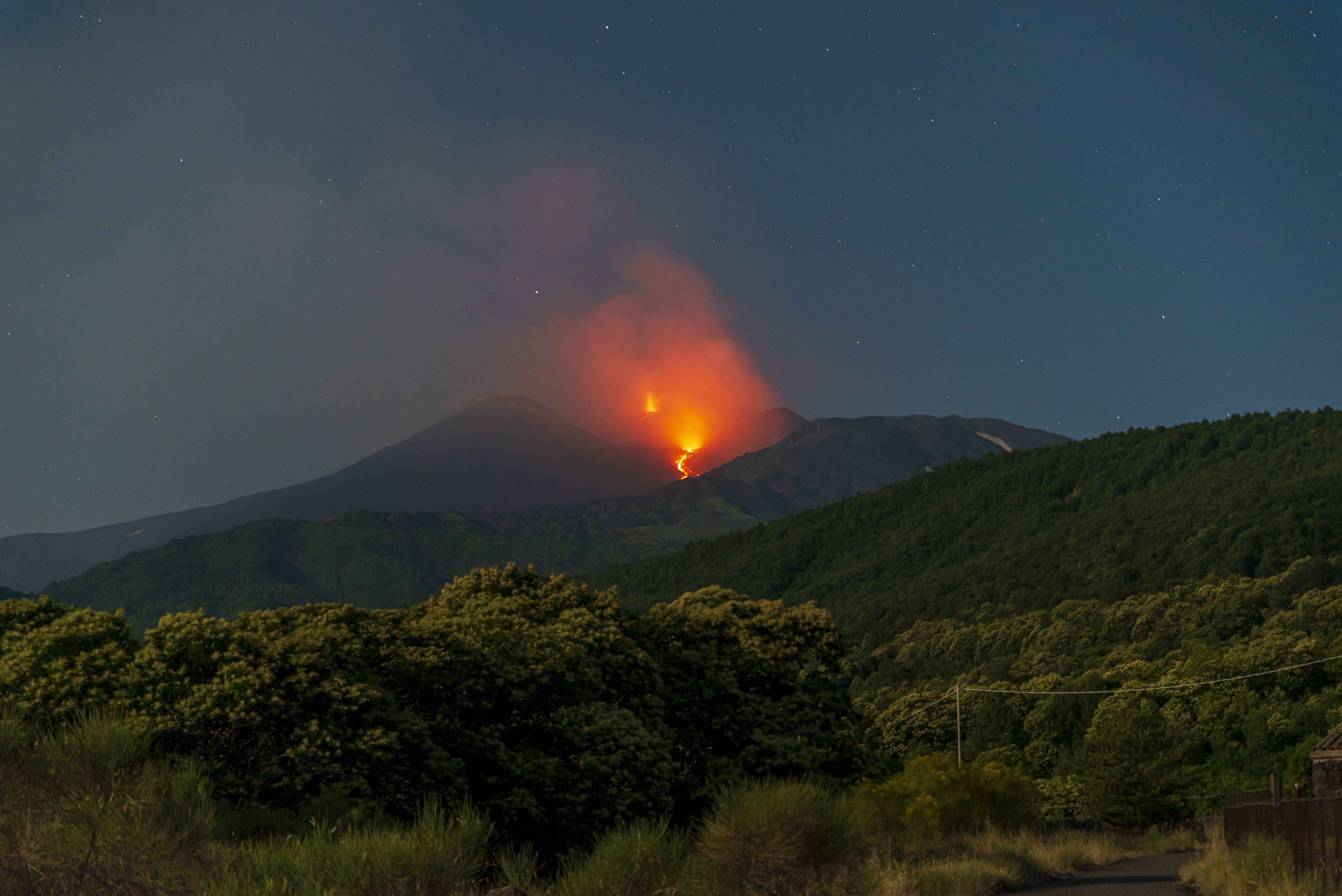 Ausbruch des Ätna bei Nacht, glühende Lava und Rauch steigen über bewaldeten Hängen auf.