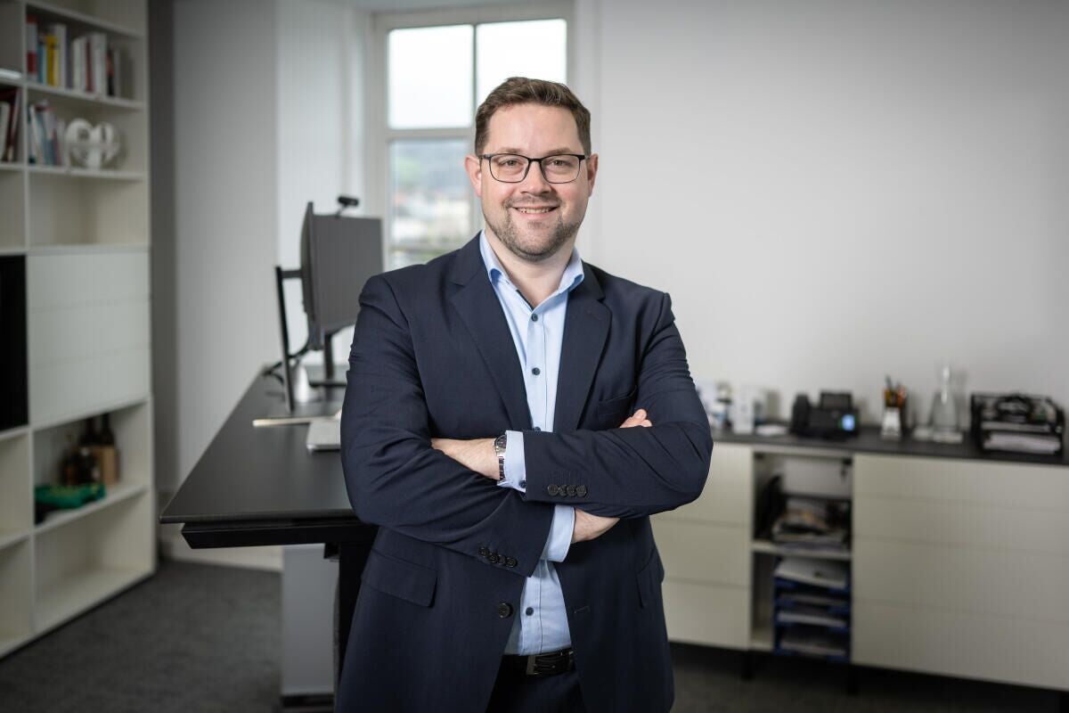 Mann im Anzug steht mit verschränkten Armen in einem modernen Büro und lächelt in die Kamera.