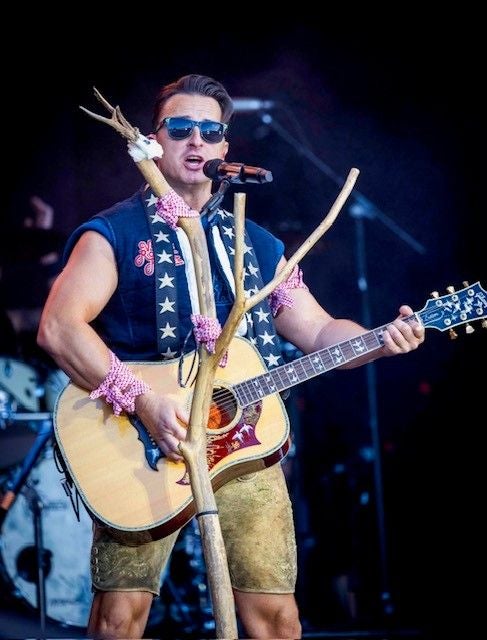Mann in Tracht spielt Gitarre und singt auf einer Bühne, Holzstock steht vor ihm.
