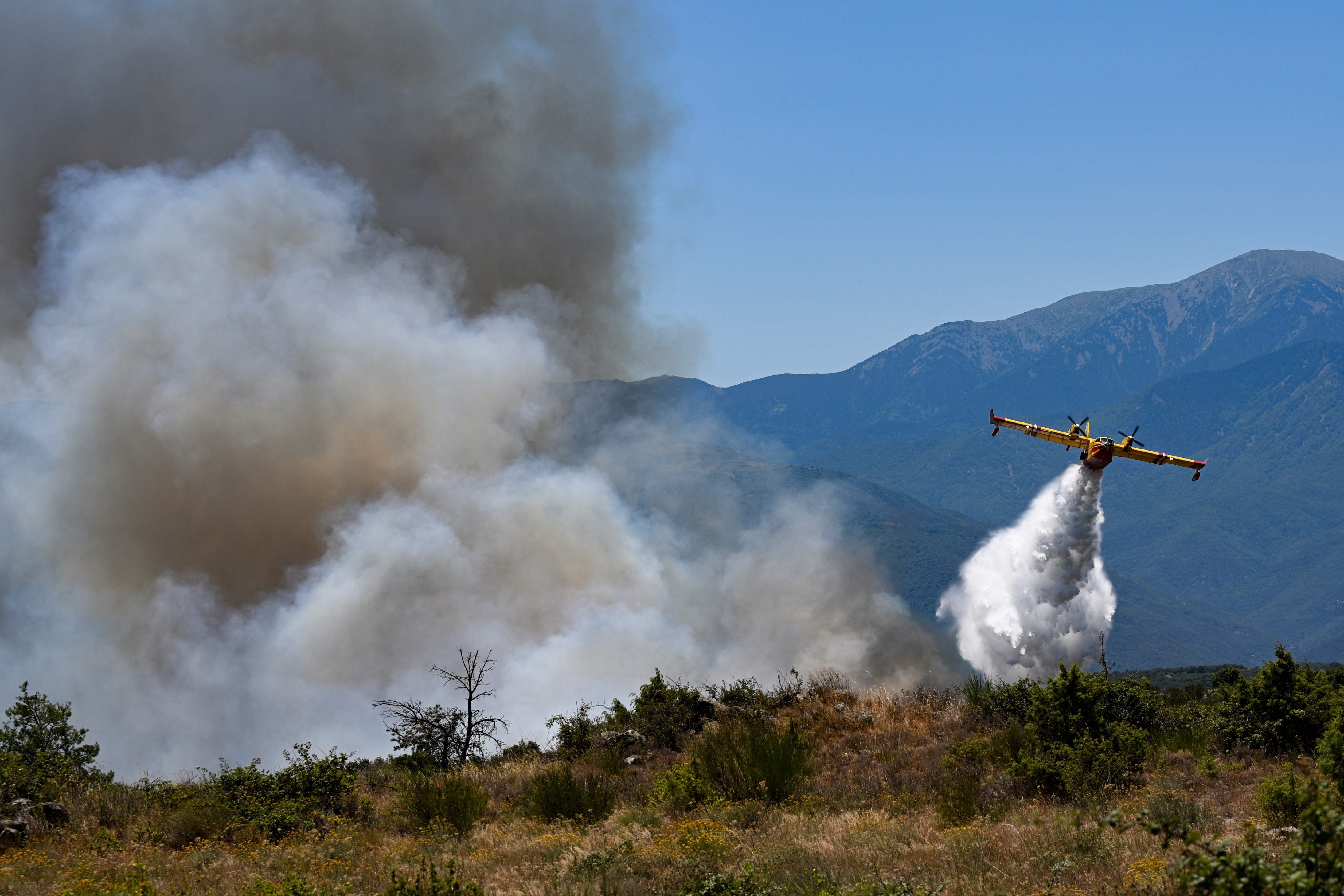 Ein Löschflugzeug wirft Wasser über einen brennenden Hang mit starkem Rauch vor Bergen ab.