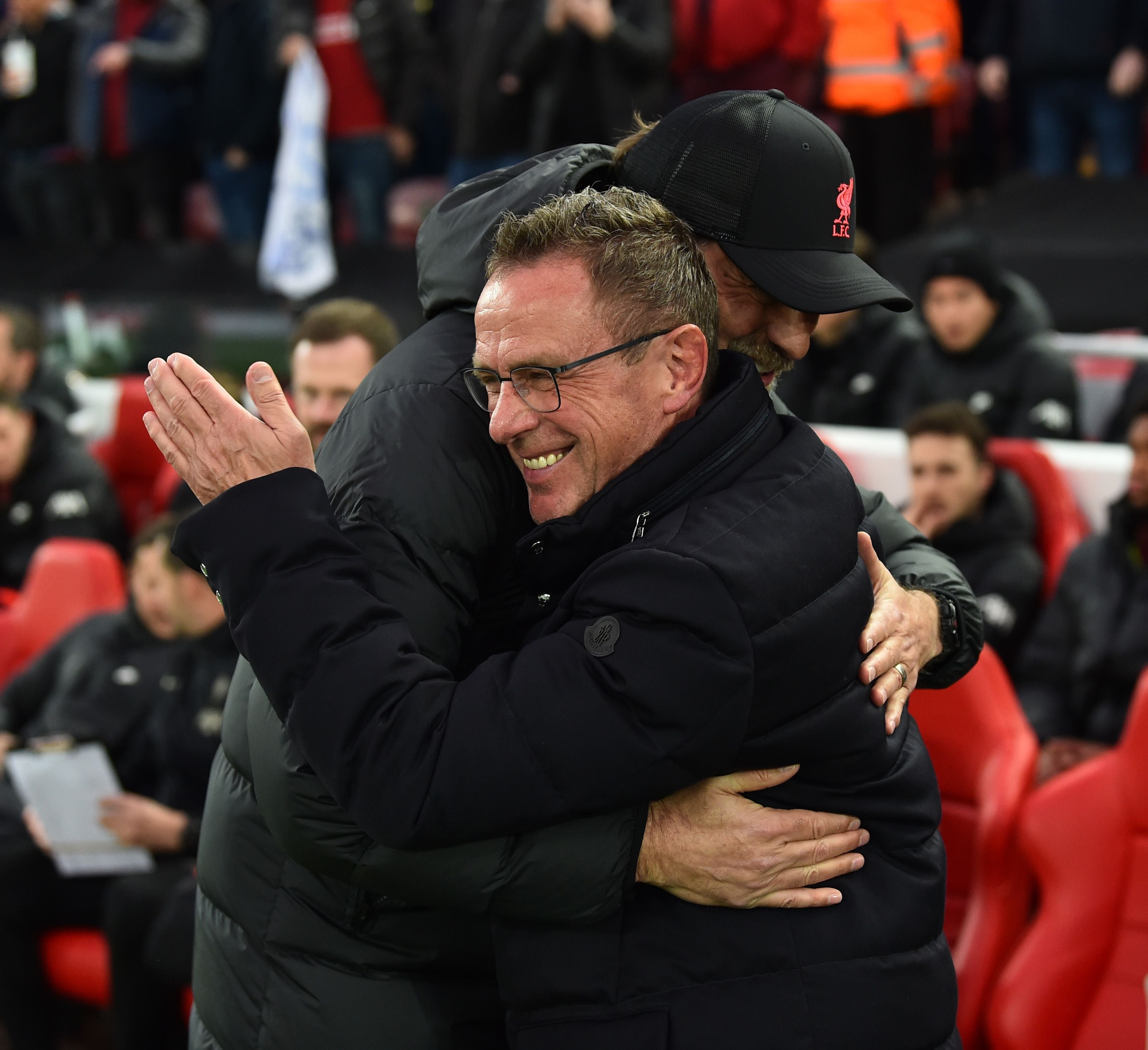 Zwei Fußballtrainer umarmen sich lächelnd am Spielfeldrand eines Stadions.