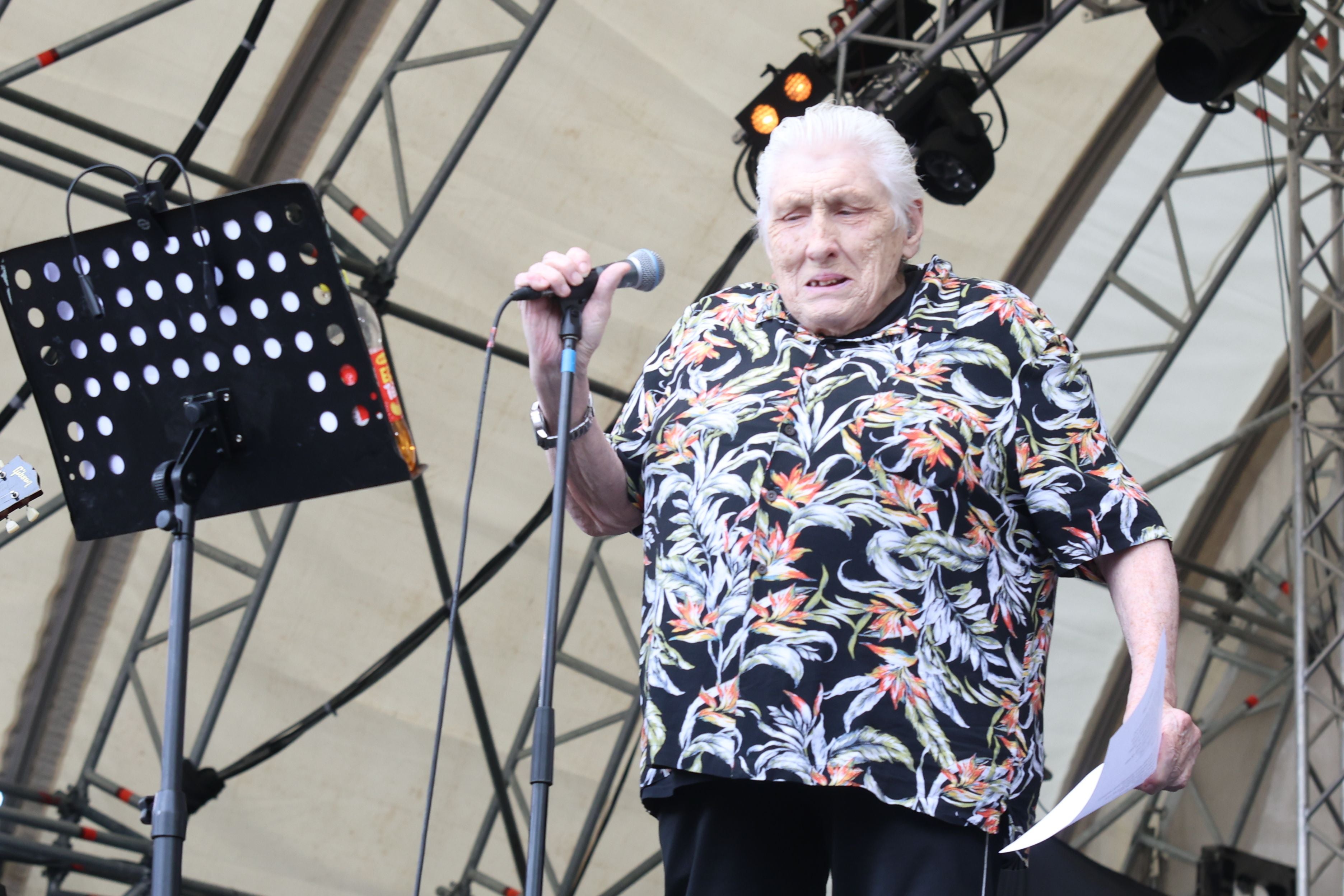 Ältere Person mit Mikrofon und Notizzettel auf einer Festivalbühne im Freien.