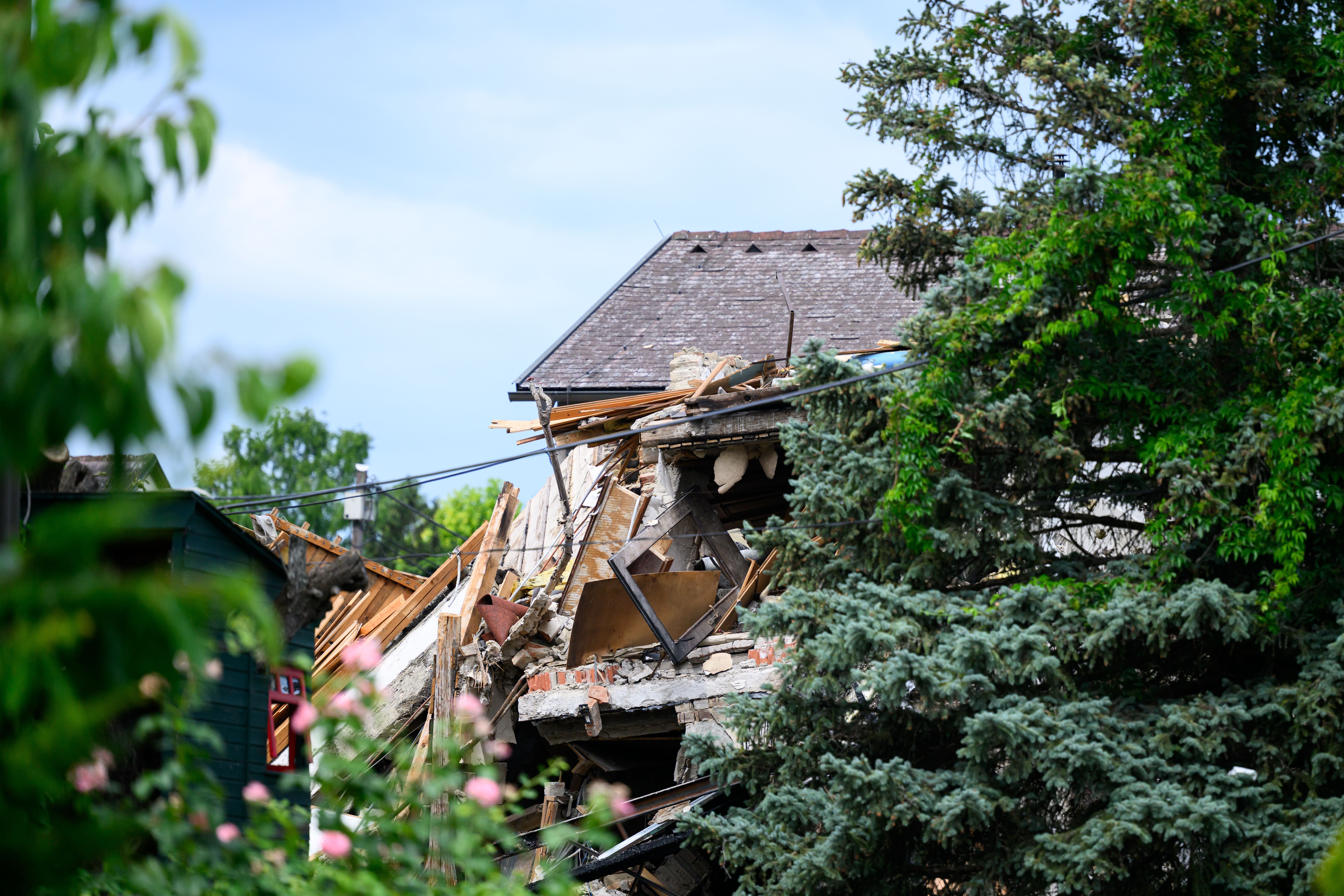 Teilweise eingestürztes Wohnhaus nach einer Explosion, umgeben von Bäumen und Pflanzen.