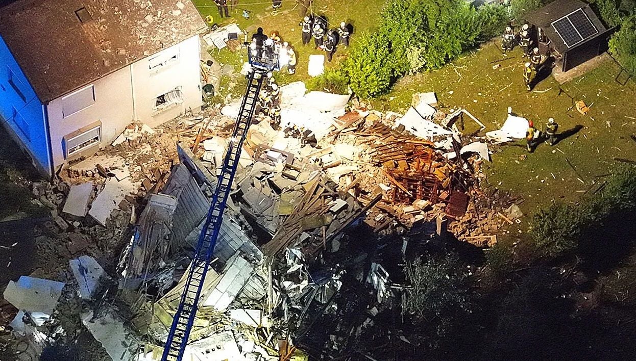Luftbild zeigt ein Haus nach einer Explosion, umgeben von Trümmern und Einsatzkräften bei Nacht.