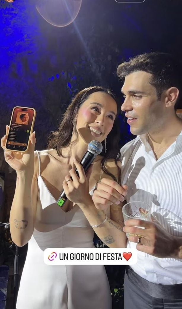 Eine Frau im weißen Kleid hält ein Mikrofon und Smartphone, ein Mann steht neben ihr.
