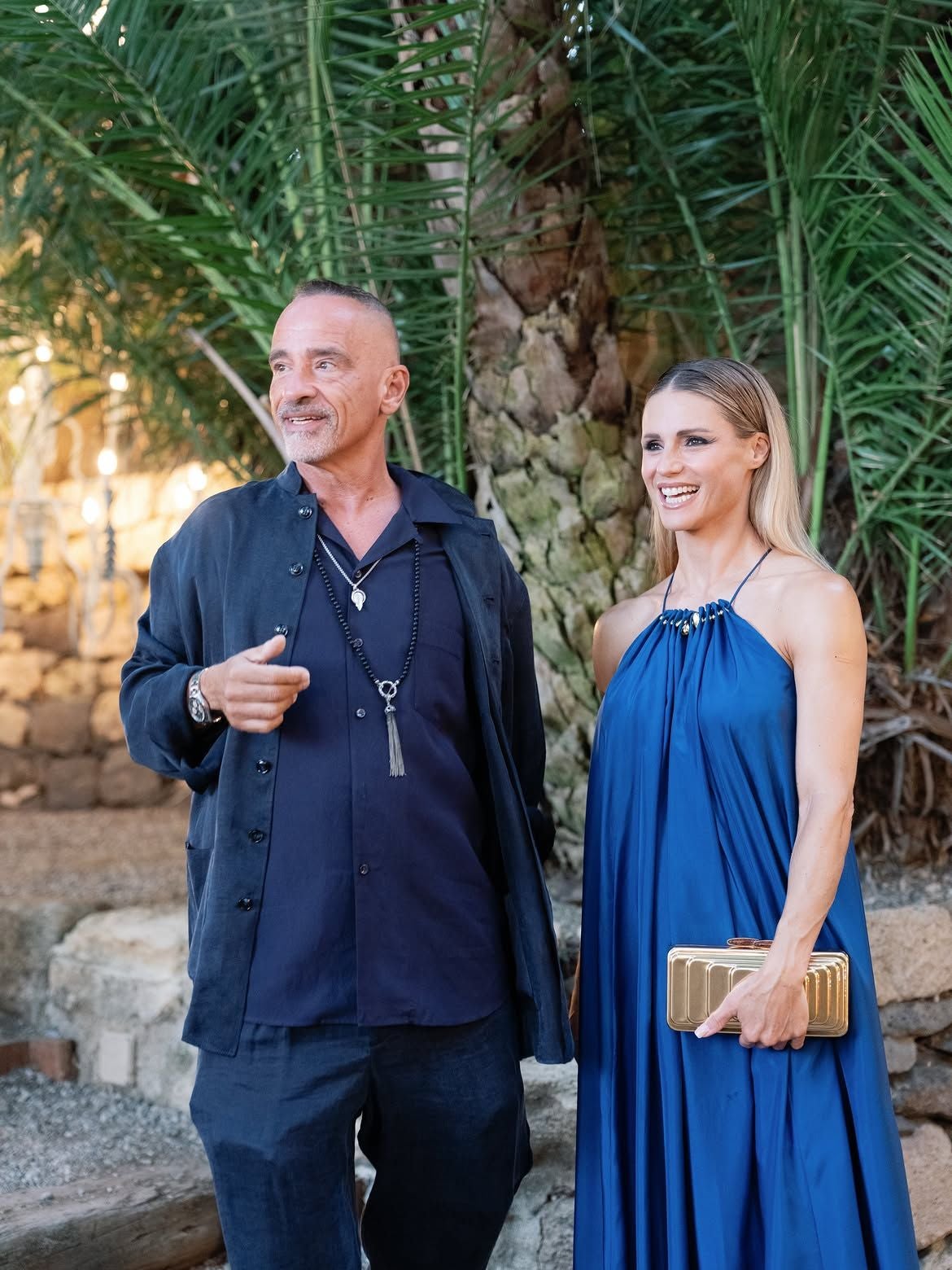 Ein Mann und eine Frau stehen elegant gekleidet vor Palmen bei einer Hochzeit.
