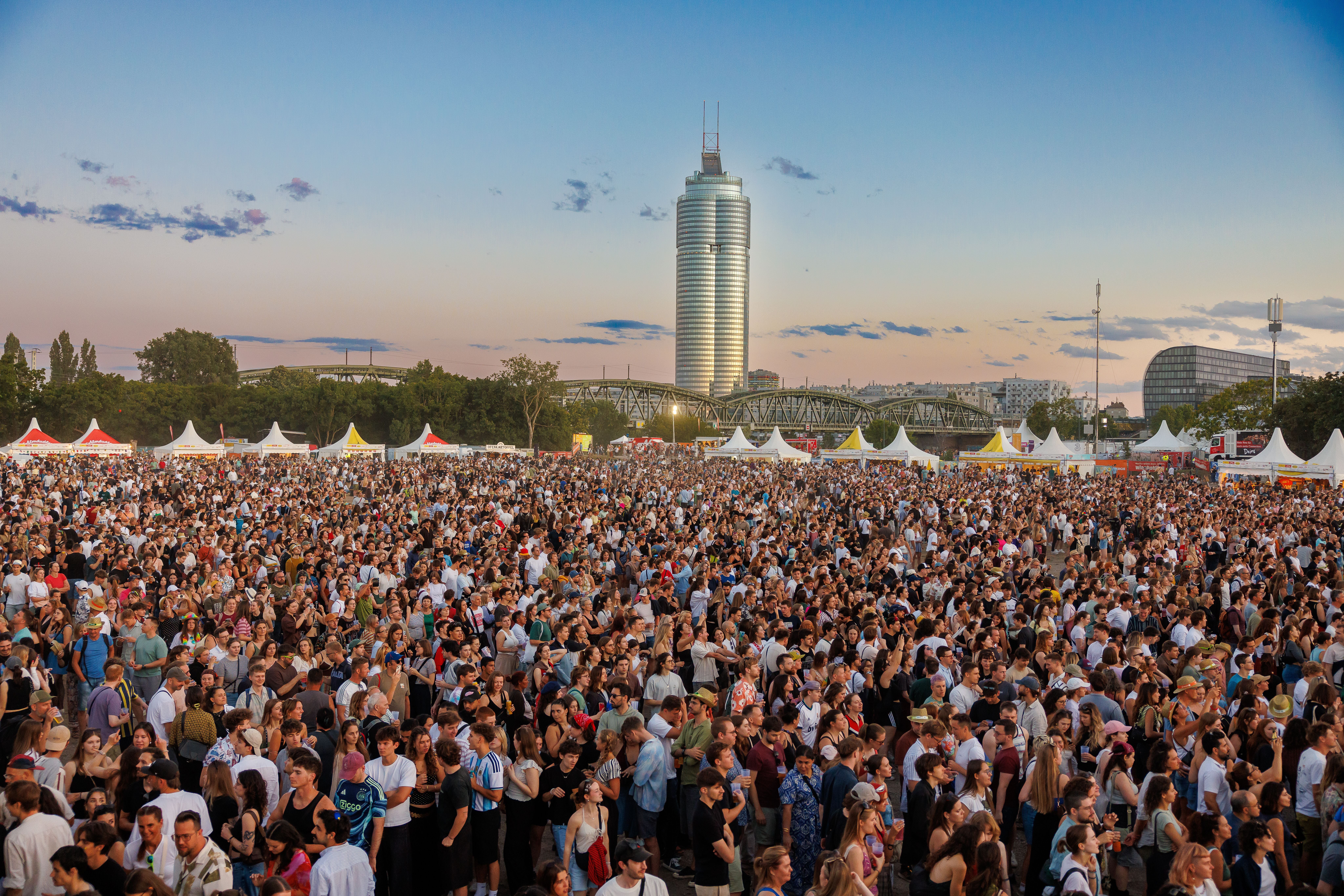 Große Menschenmenge auf dem Donauinselfest in Wien mit Zeltständen und Hochhaus im Hintergrund.