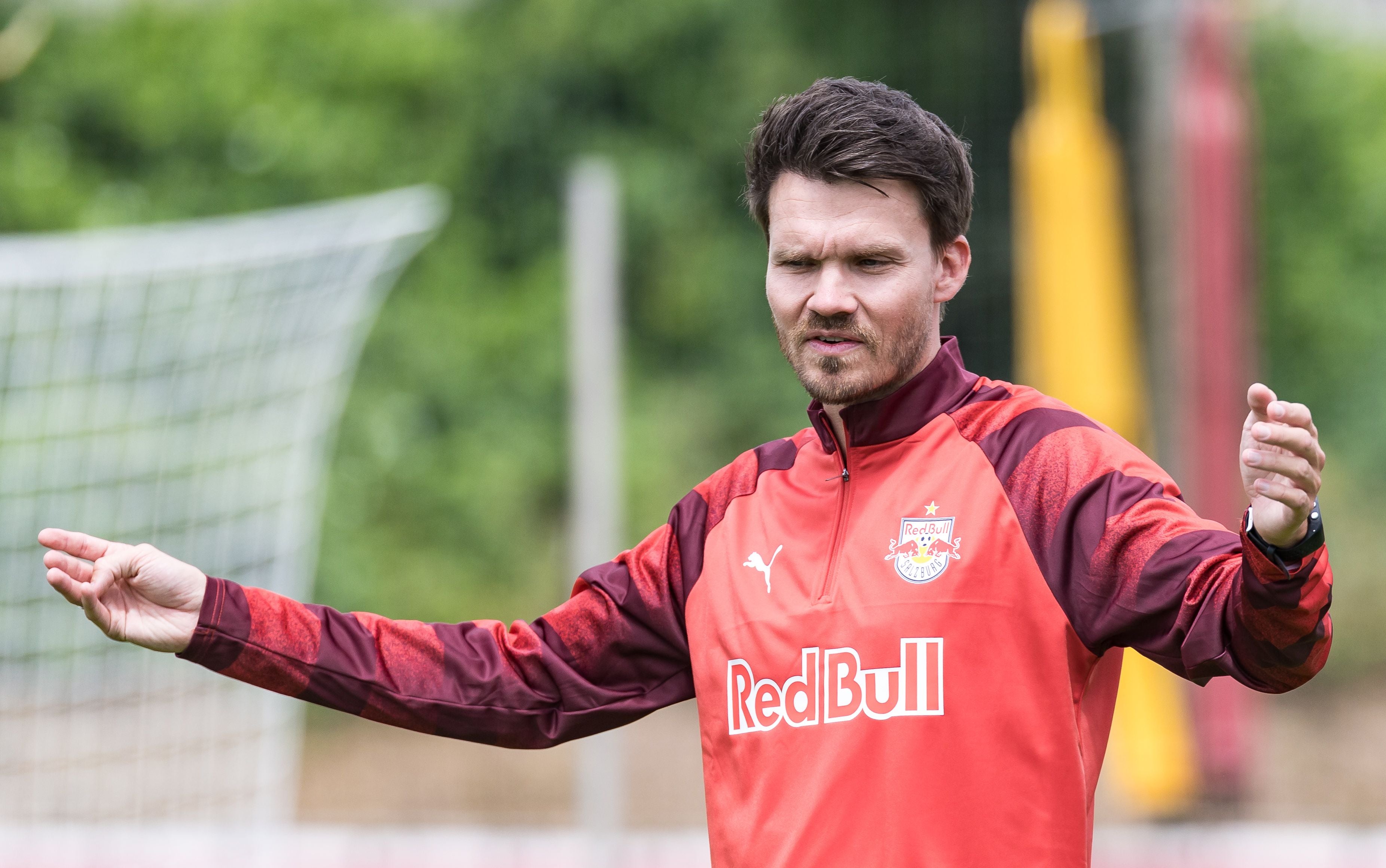 Ein Trainer im Red Bull Salzburg Trainingsanzug gestikuliert auf einem Fußballplatz.