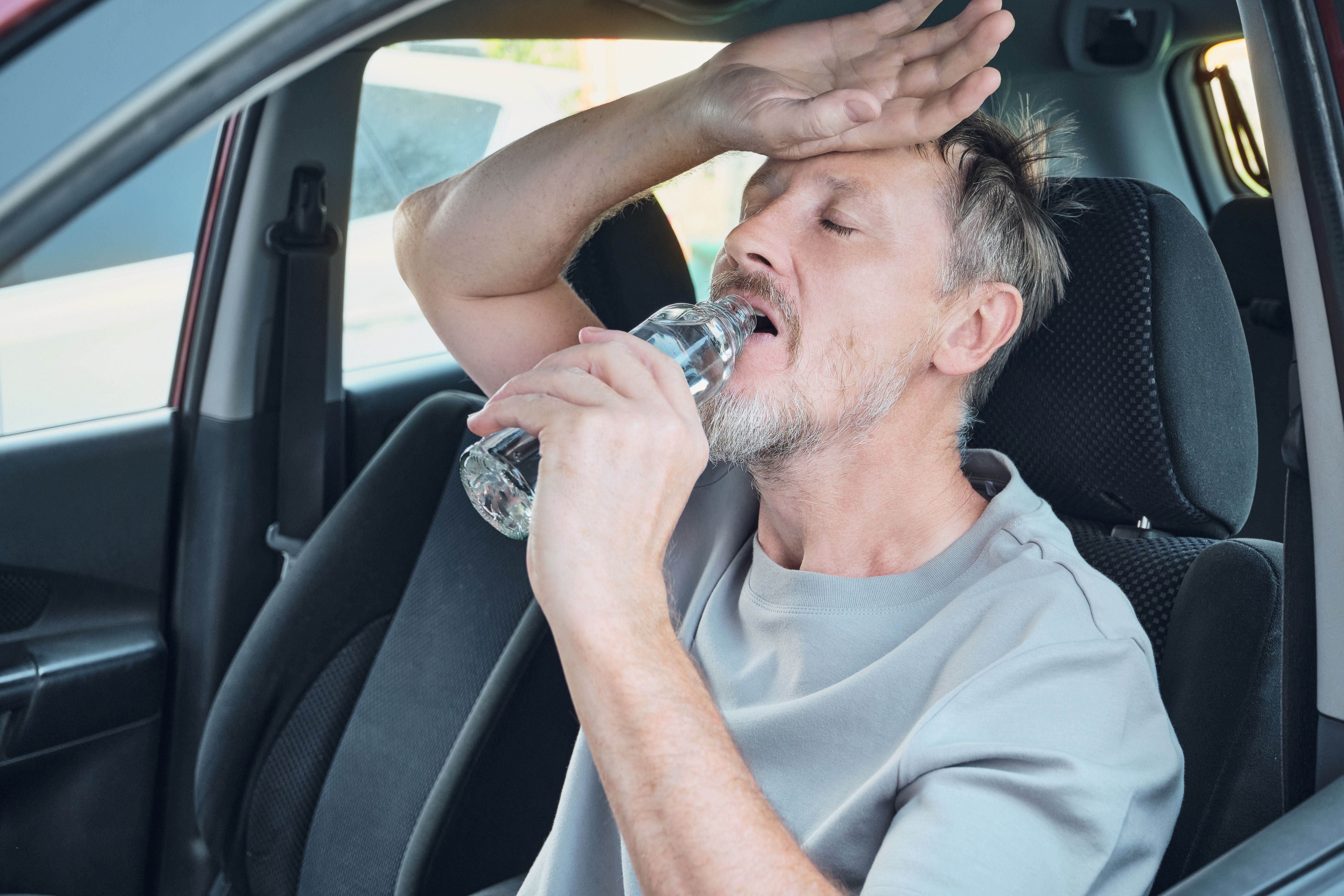 Mann trinkt im Auto Wasser, wirkt erschöpft und schwitzt vor Hitze.