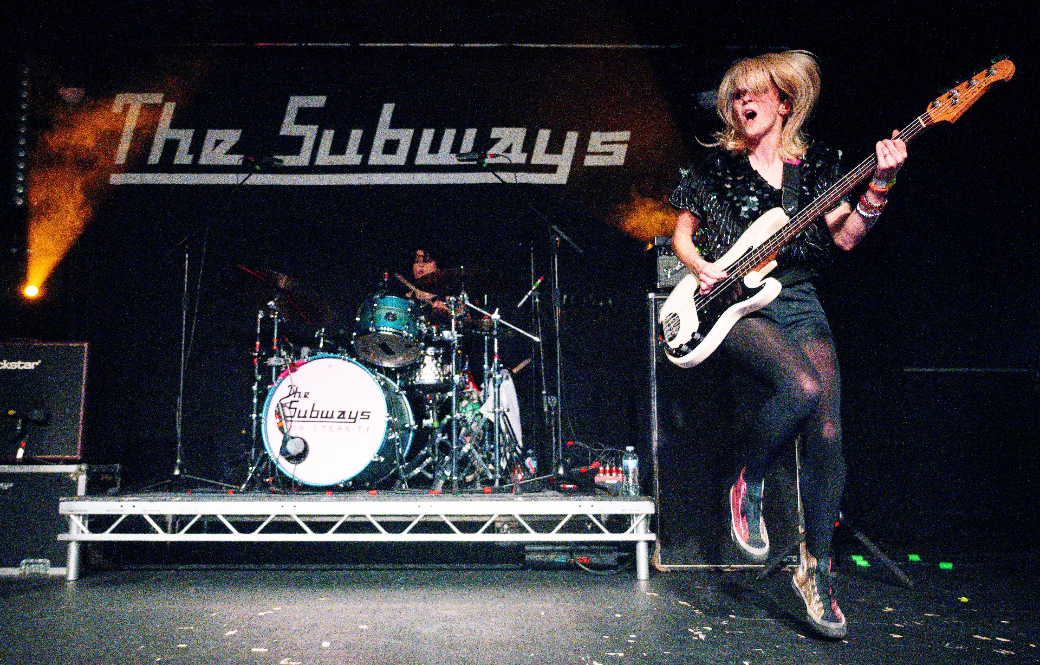 Bassist springt mit Energie auf der Bühne, Schlagzeugerin spielt dahinter, „The Subways“-Logo im Hintergrund.