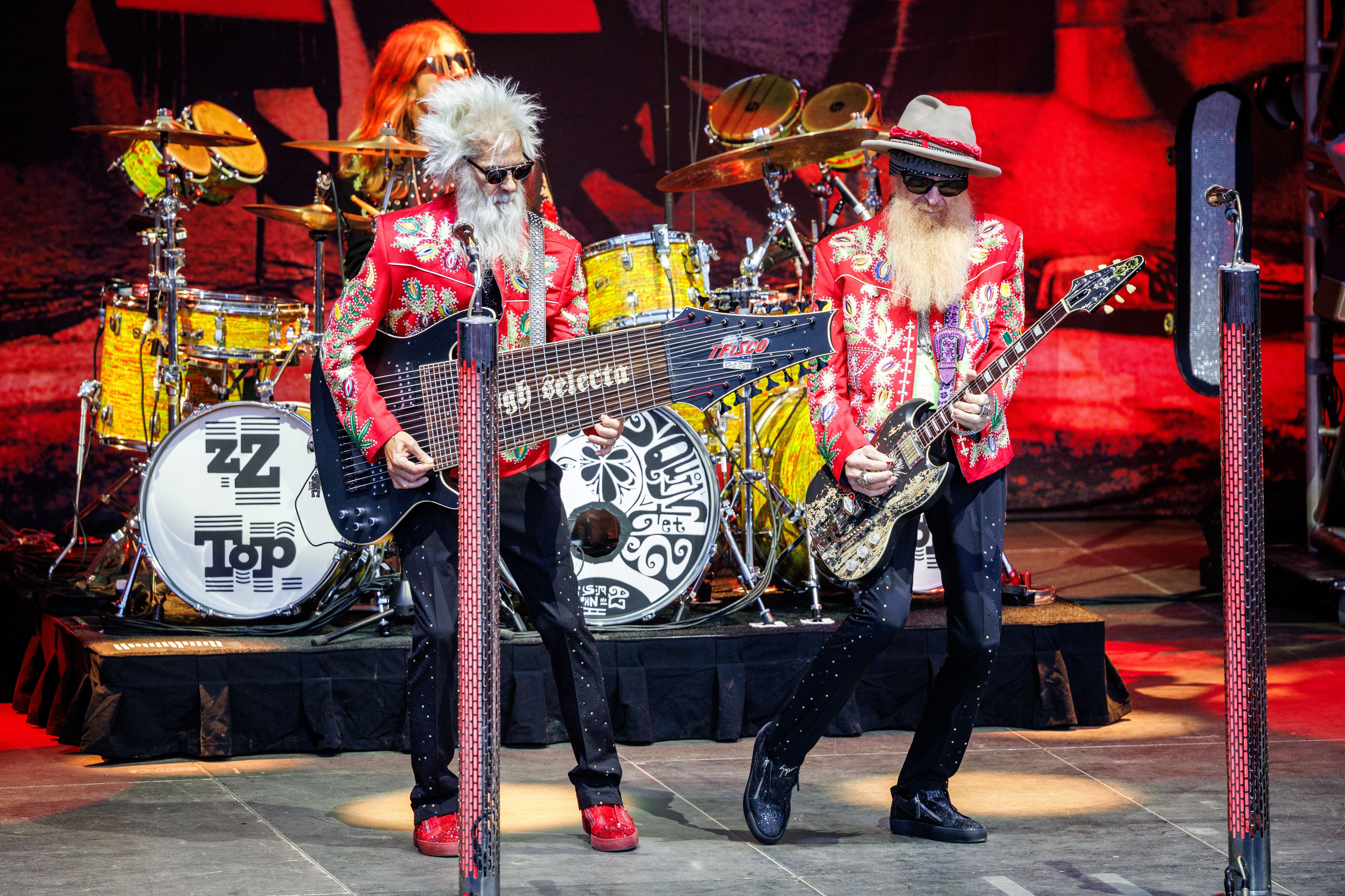 Drei Mitglieder der Band ZZ Top spielen in bunten Jacken auf einer Bühne mit Schlagzeug im Hintergrund.