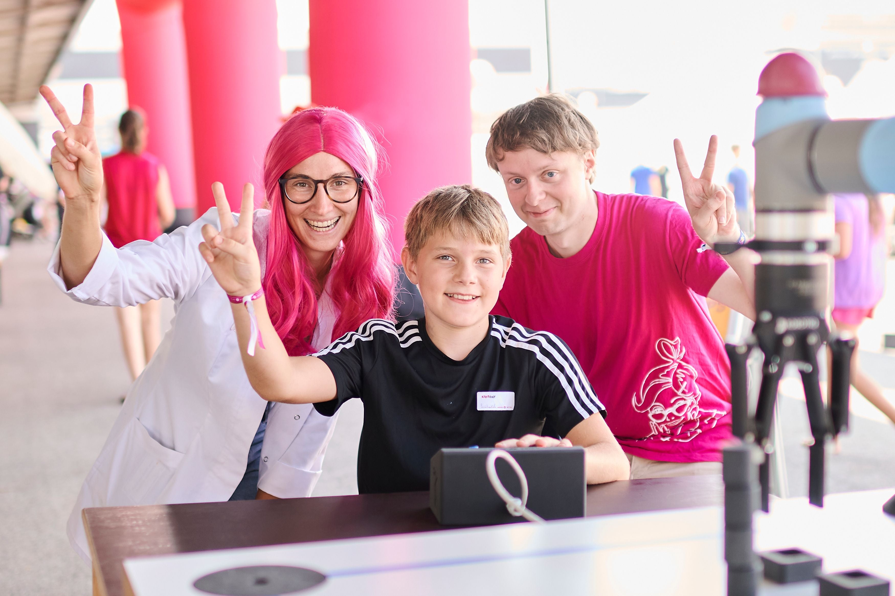 Drei lachende Personen beim Ferienspiel Wiener Netze zeigen das Victory-Zeichen an einem Tisch.