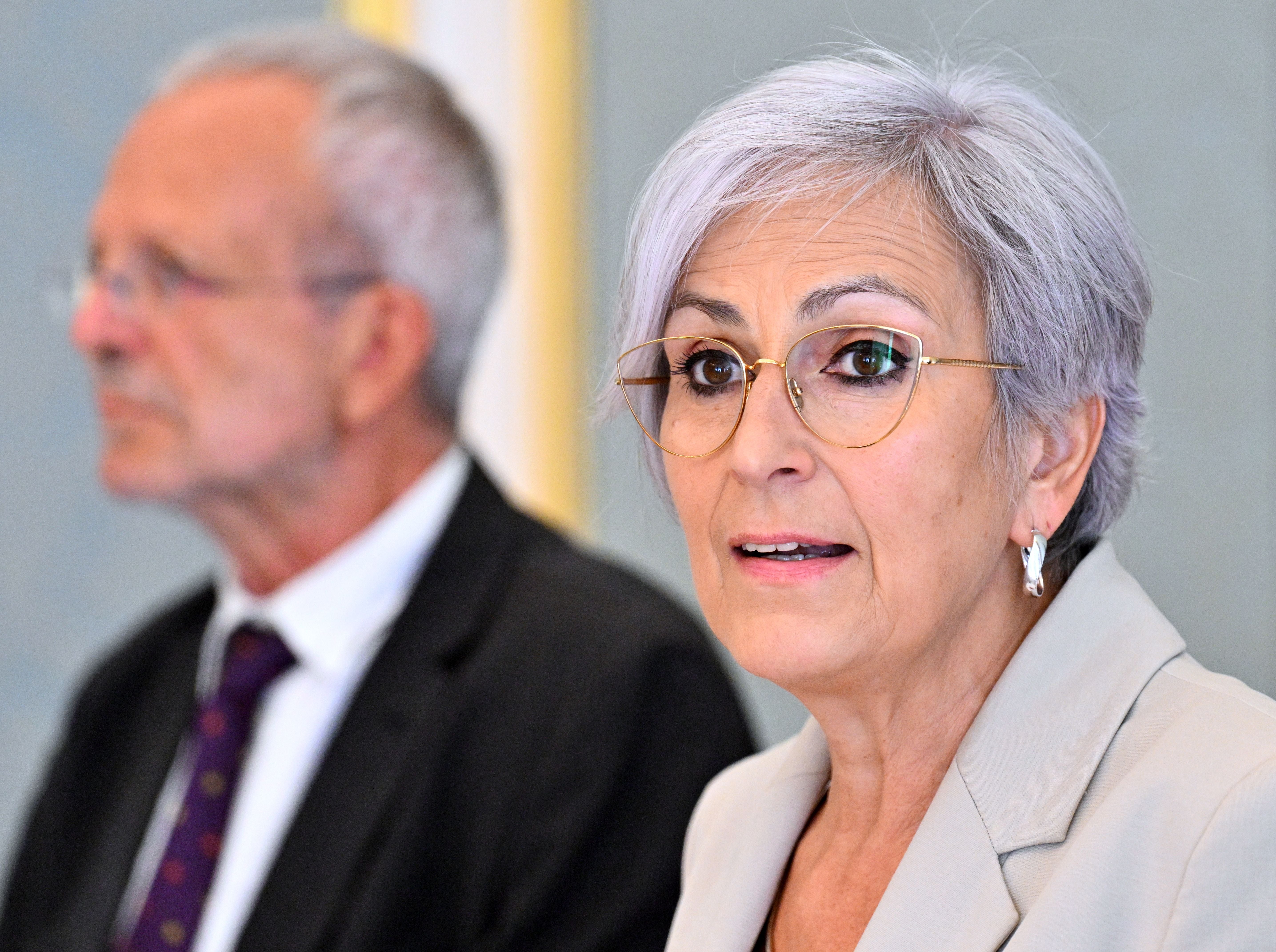 Frau mit grauen Haaren und Brille spricht, Mann im Anzug sitzt unscharf daneben.