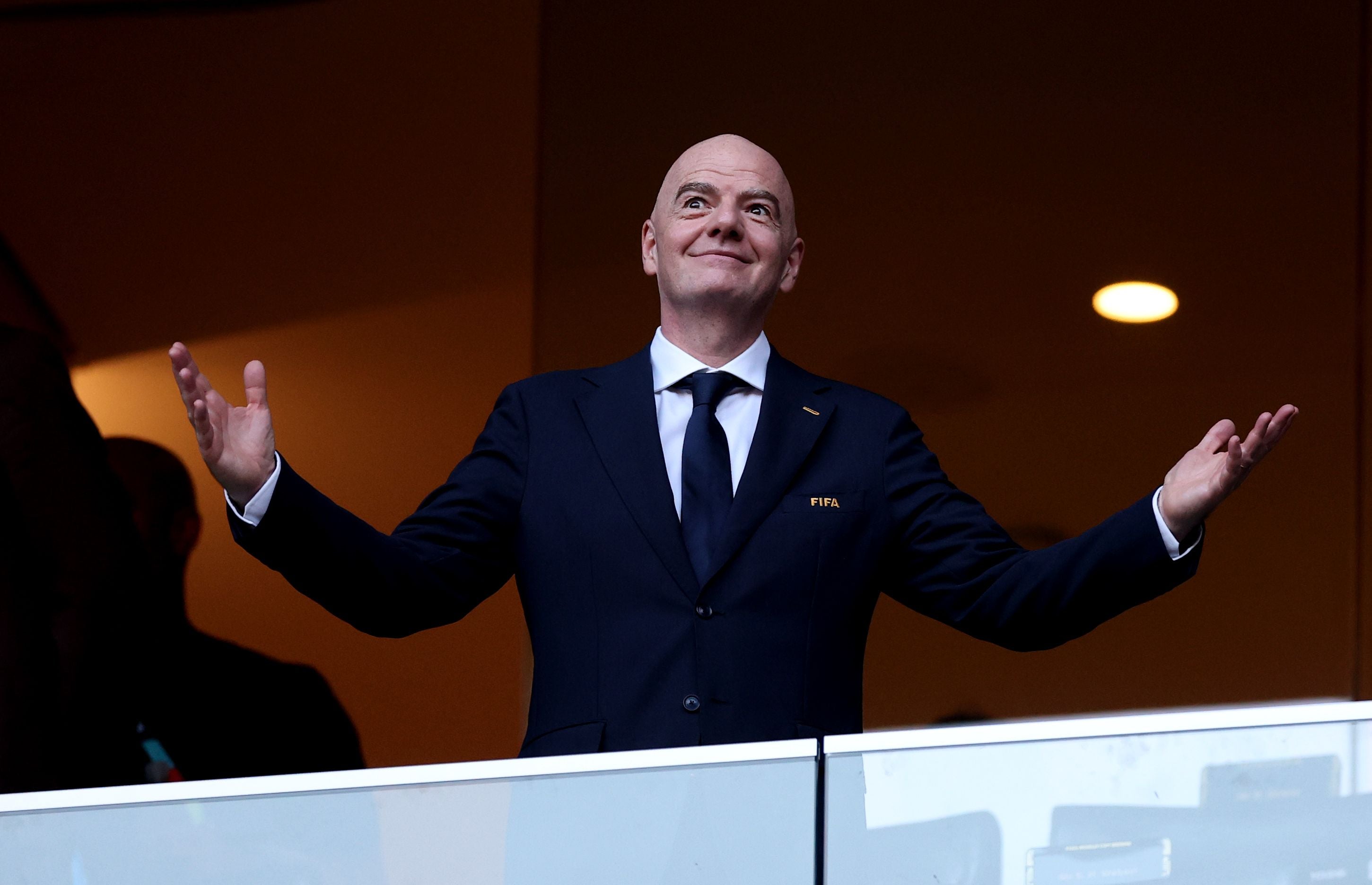 Mann im Anzug steht mit ausgebreiteten Armen und lächelt in einer Loge im Stadion.