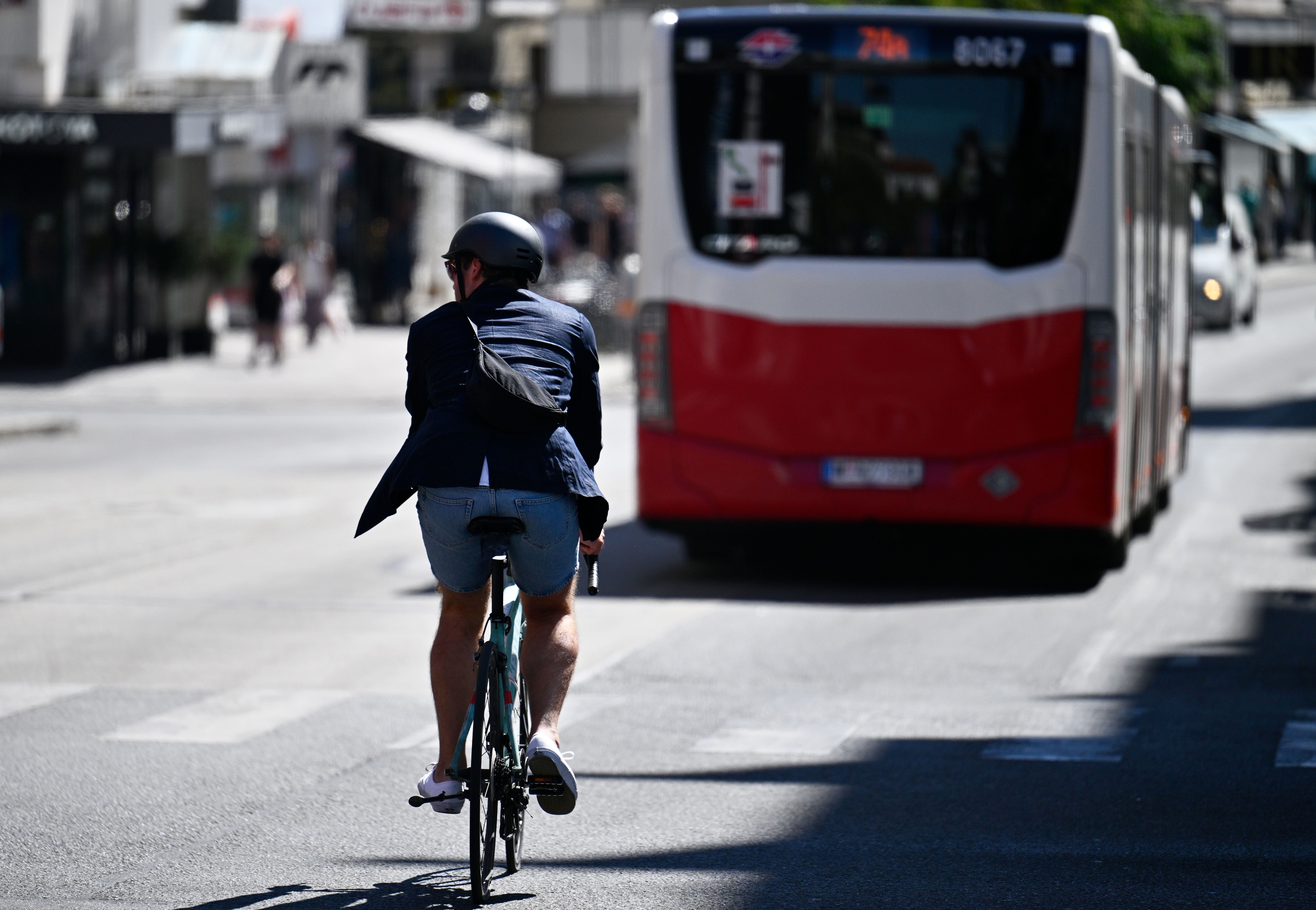 Ein Radfahrer fährt hinter einem roten Wiener Linien Bus auf einer sonnigen Stadtstraße.
