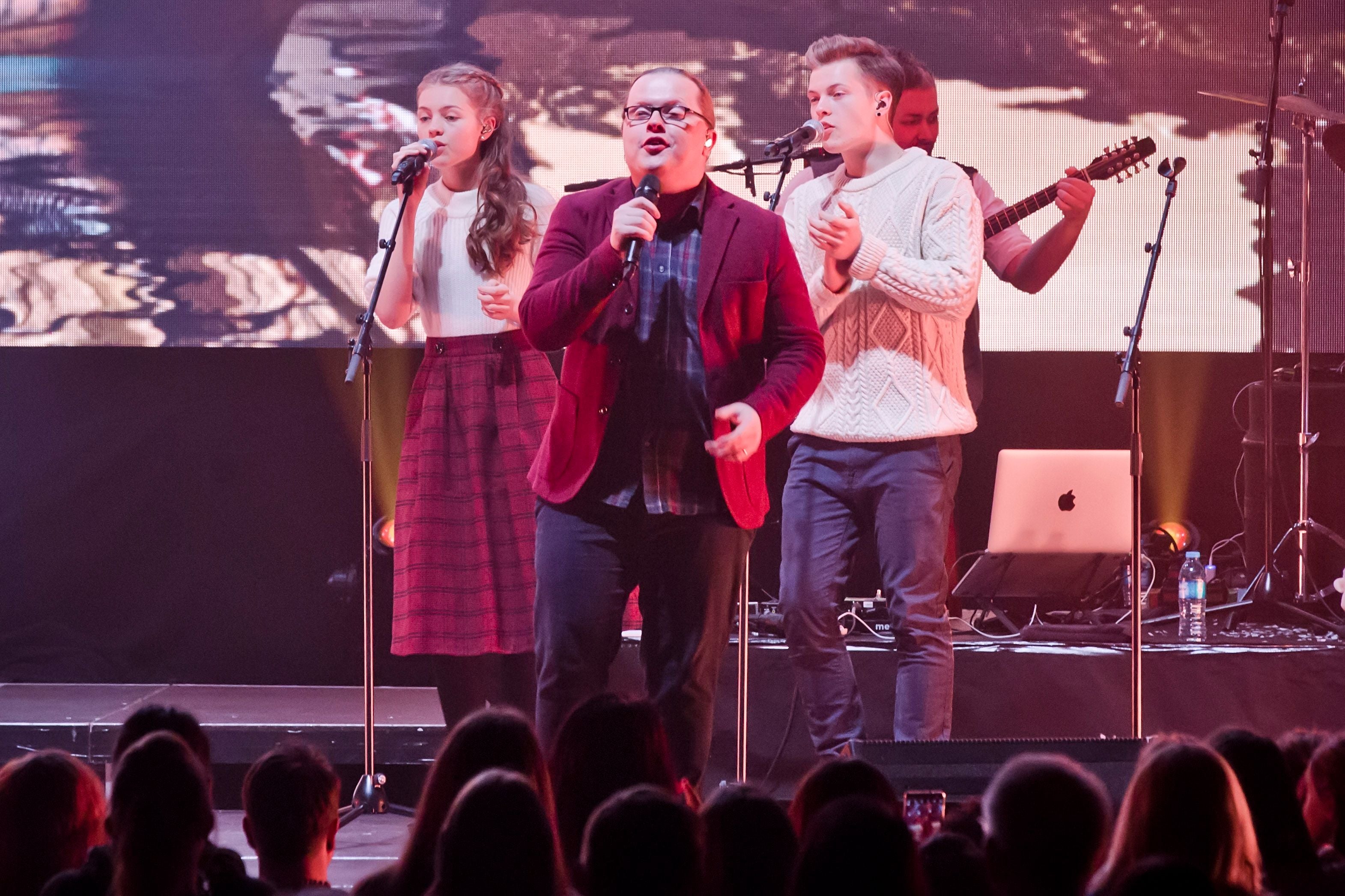 Drei Musiker singen und spielen bei einem Live-Konzert auf einer Bühne vor Publikum.