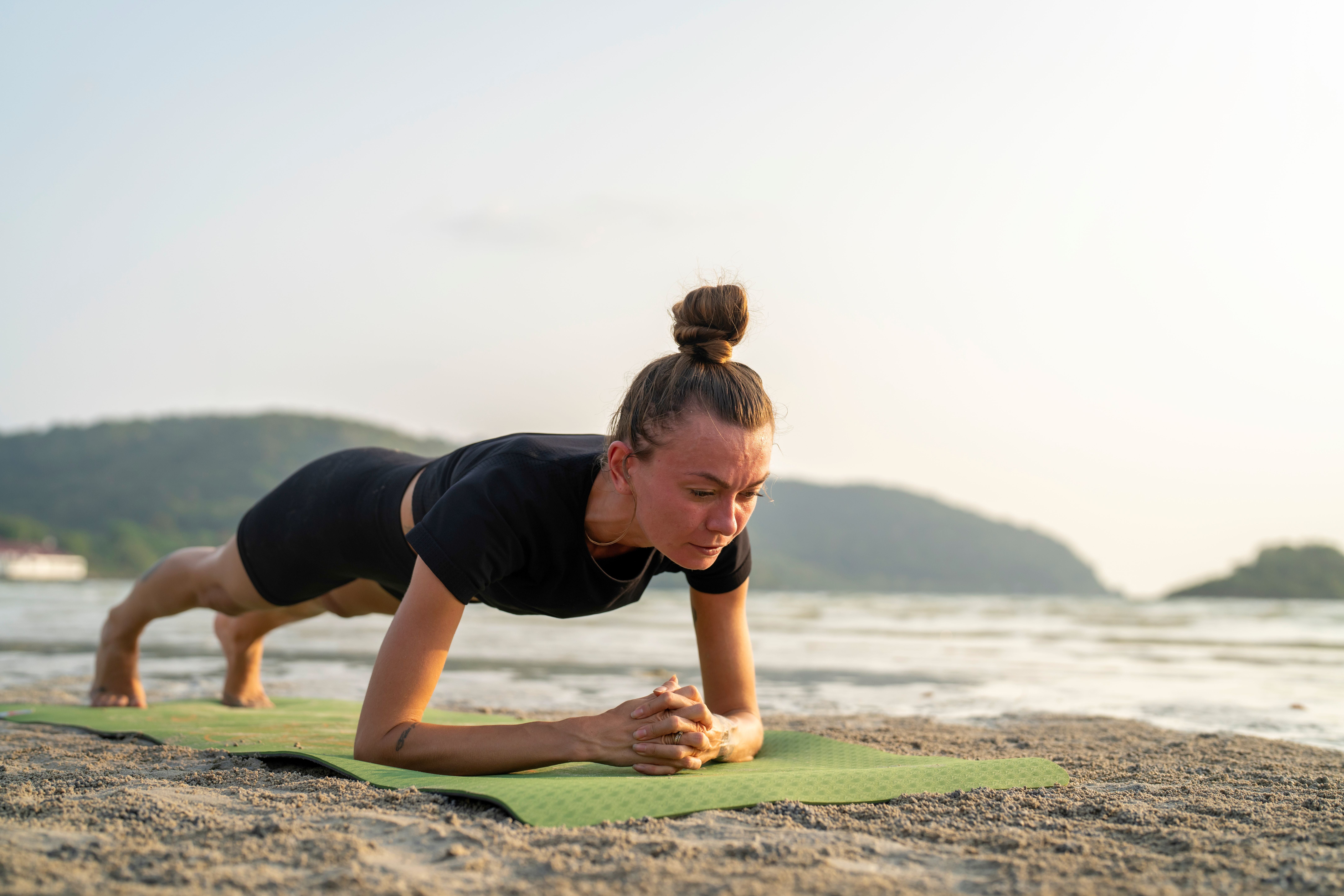 Frau macht Plank-Übung auf einer Matte am Sandstrand vor hügeliger Kulisse.
