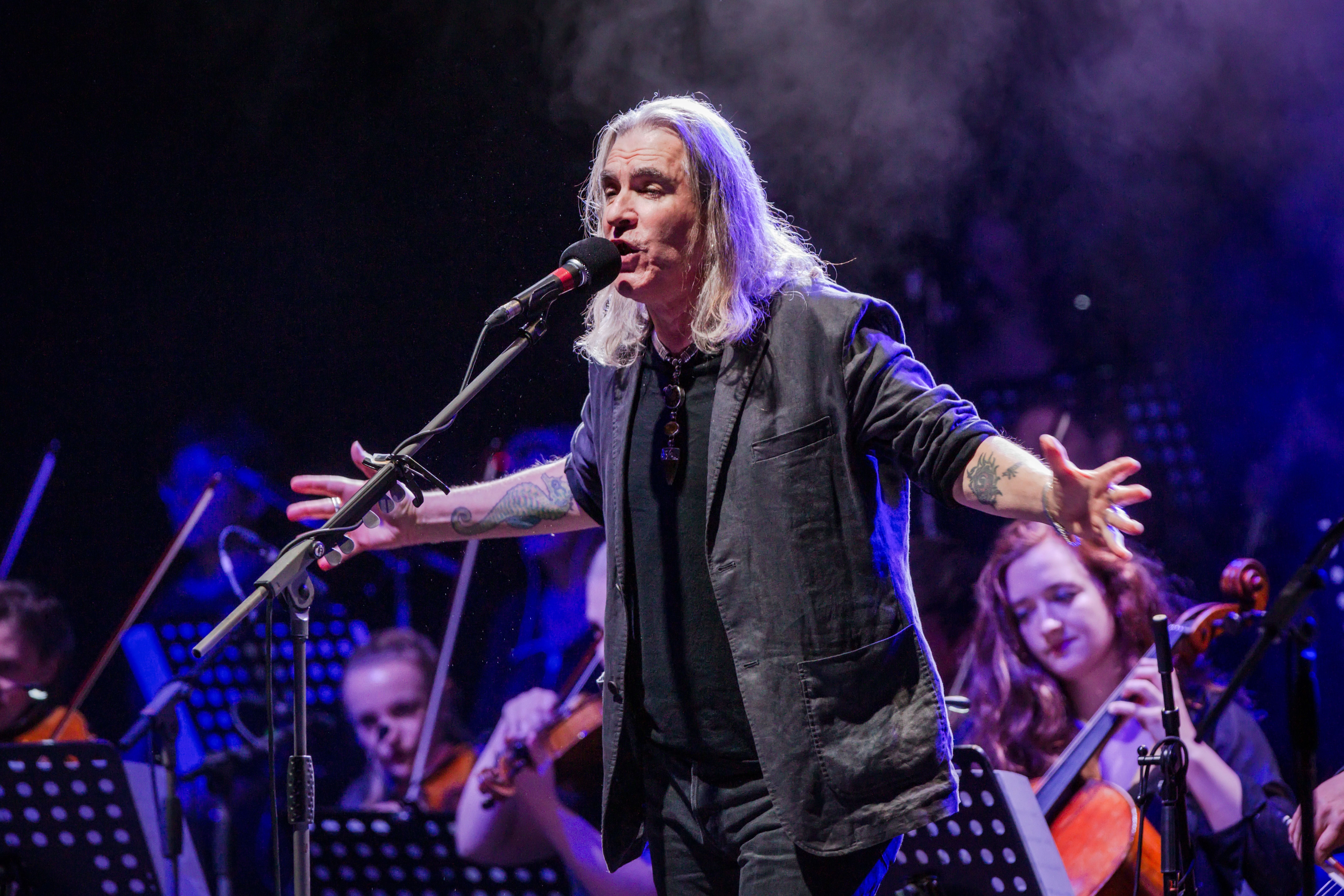 Ein Sänger mit langen grauen Haaren singt vor einem Orchester bei einem Konzert.