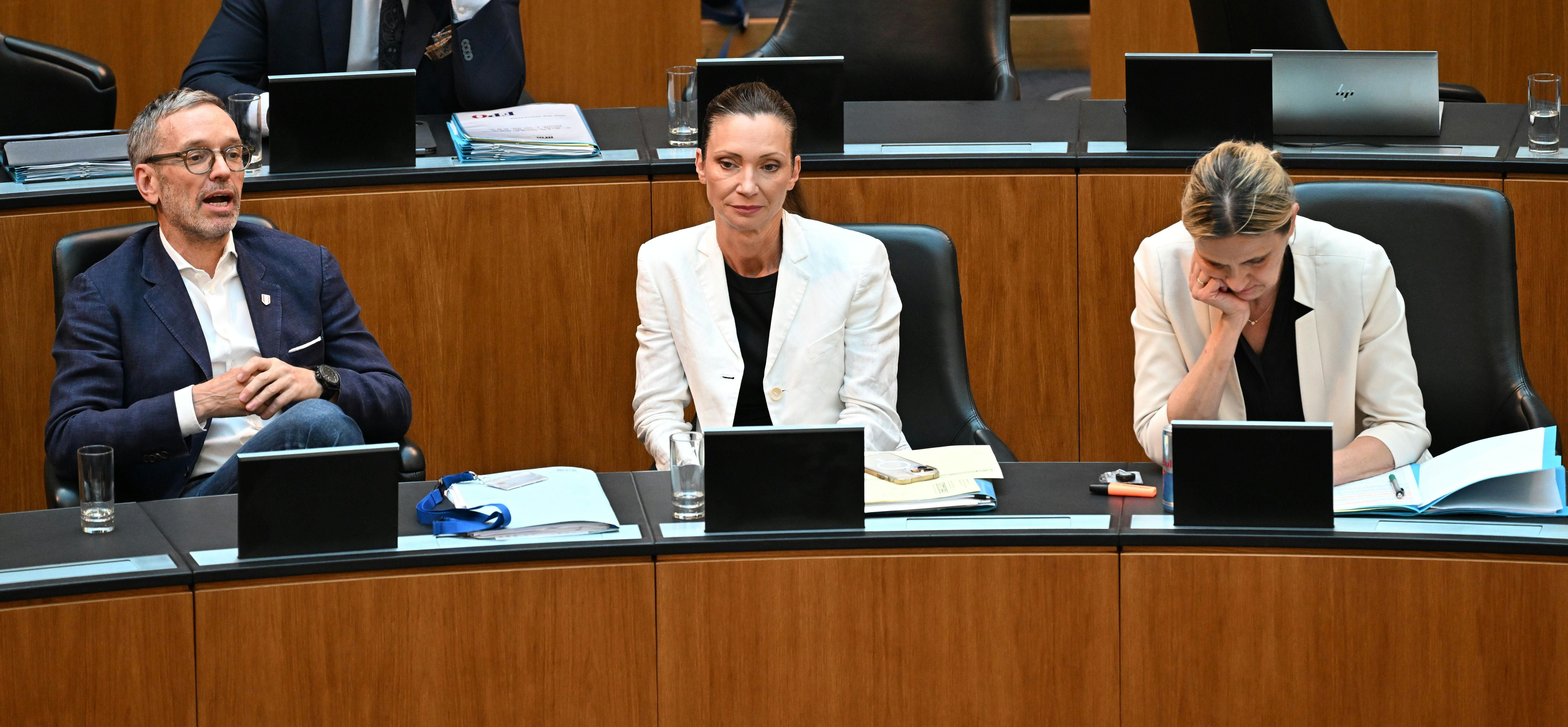 Drei Personen sitzen bei einer Nationalratssitzung an Tischen mit Laptops und Unterlagen.