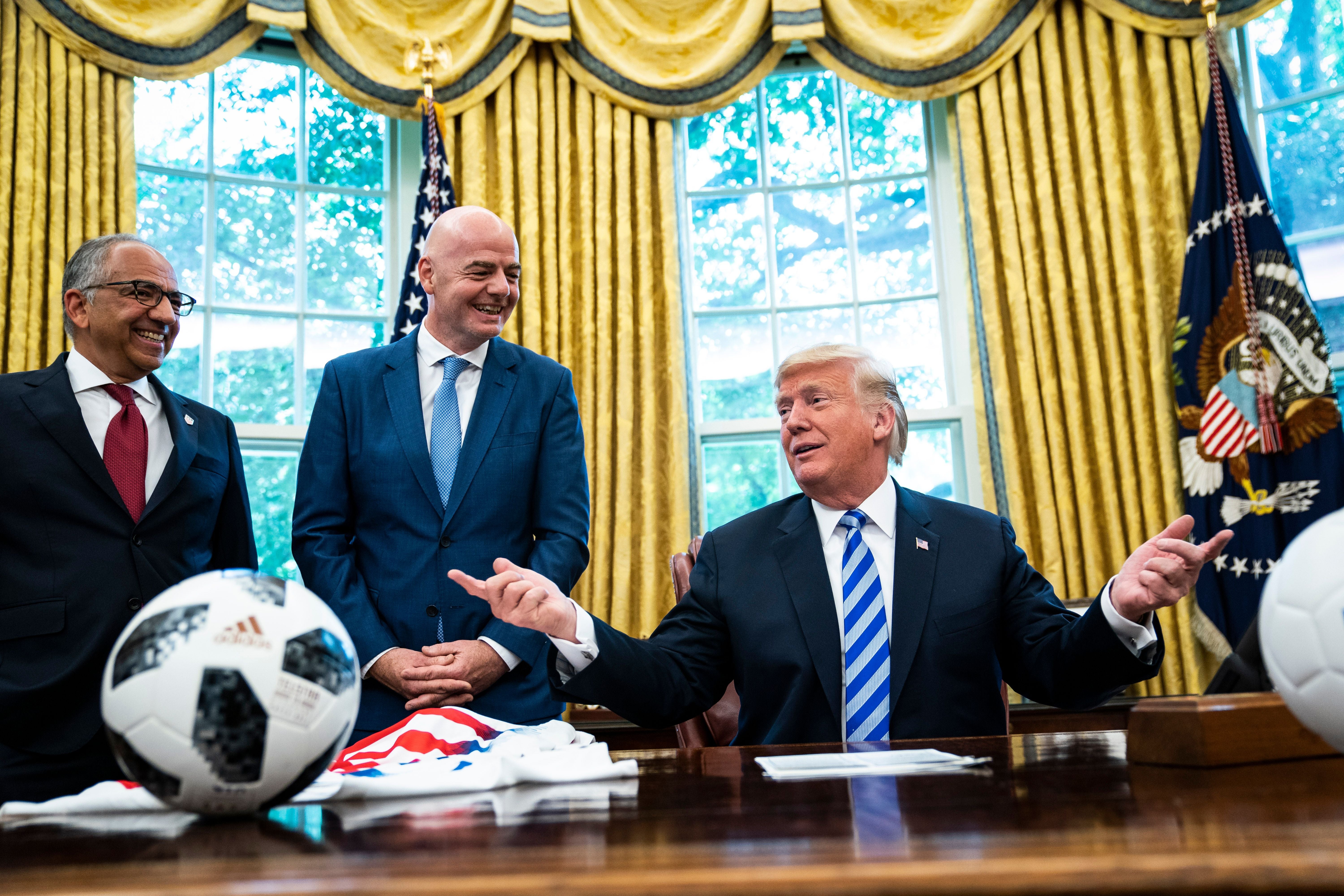 Drei Männer im Anzug sprechen im Oval Office, Fußballbälle und Trikots liegen auf dem Tisch.