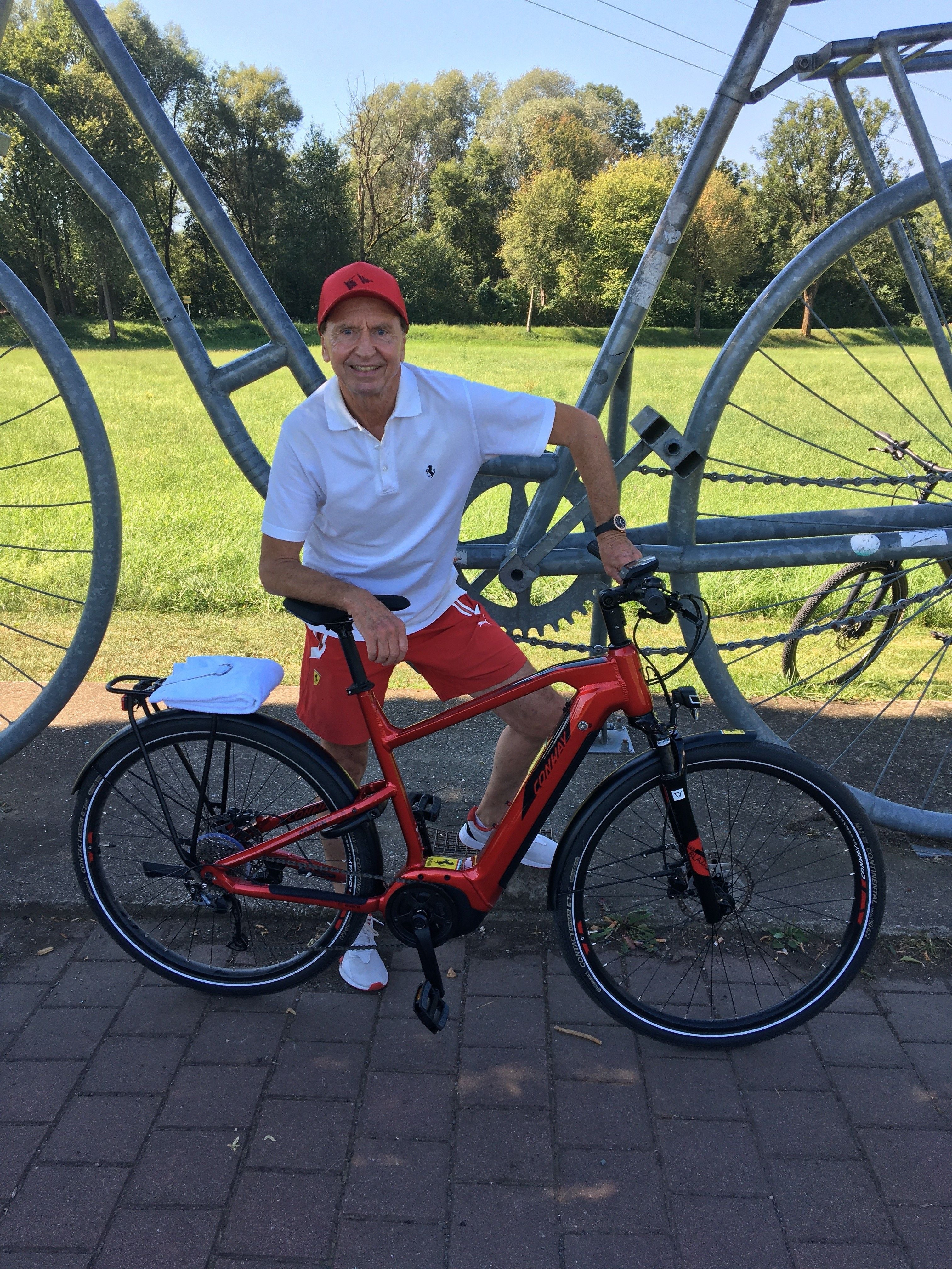 Ein Mann im Sportoutfit posiert mit rotem Fahrrad vor einer großen Fahrradskulptur im Park.