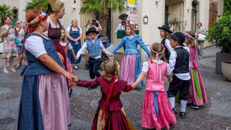Kinder in bunten Trachten tanzen im Kreis auf einer gepflasterten Straße bei einem Fest.