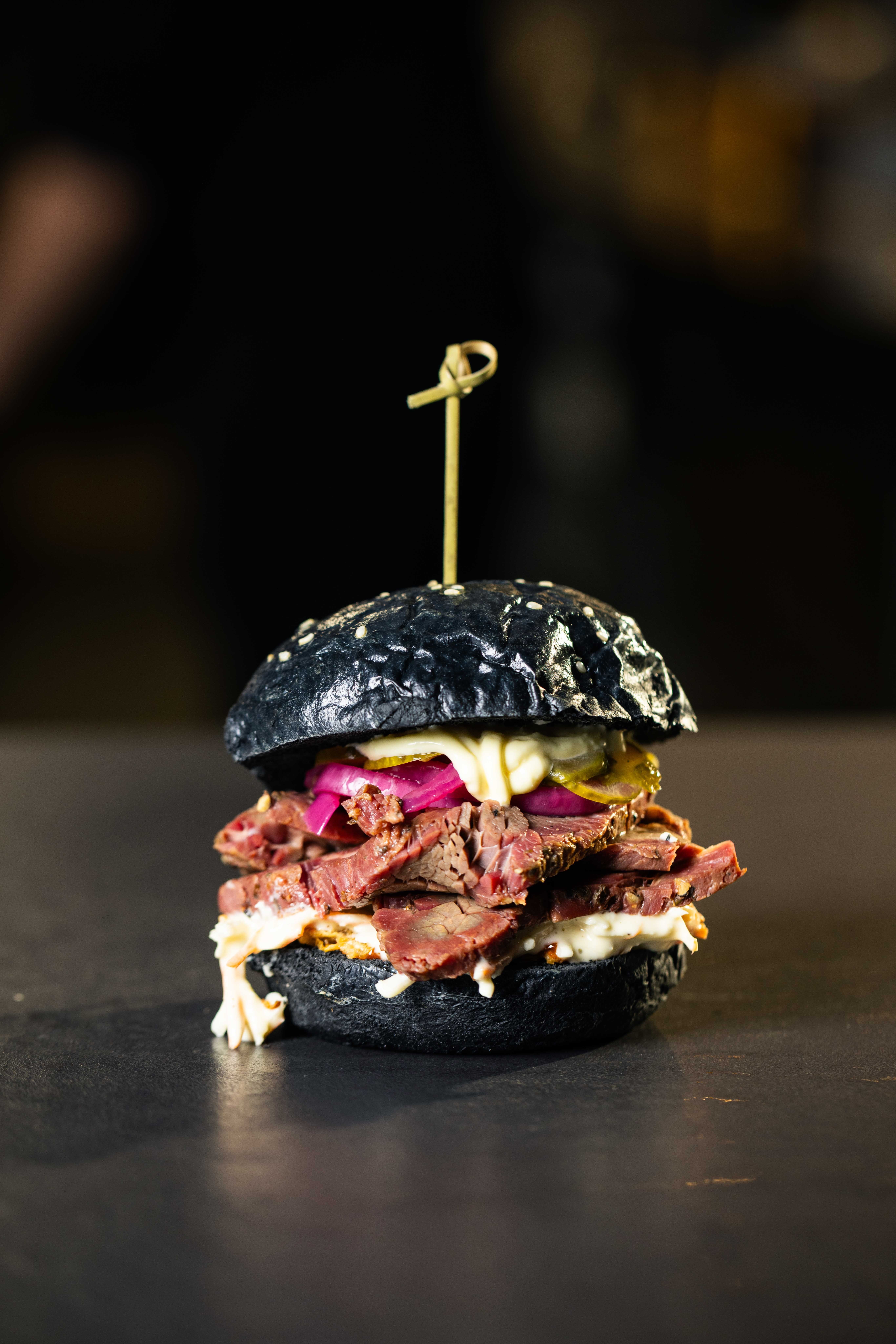 Schwarzer Burger mit Rindfleisch, Zwiebeln, Gurken und Soße auf dunklem Hintergrund.
