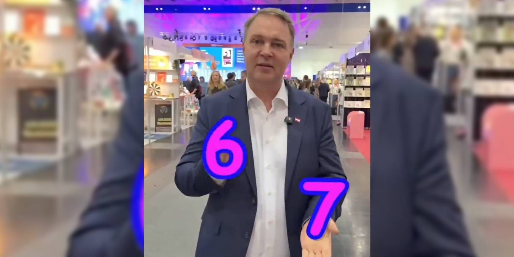 Ein Mann im Anzug hält die Zahlen 6 und 7, digital eingefügt, auf seinen Händen in einer Messehalle.