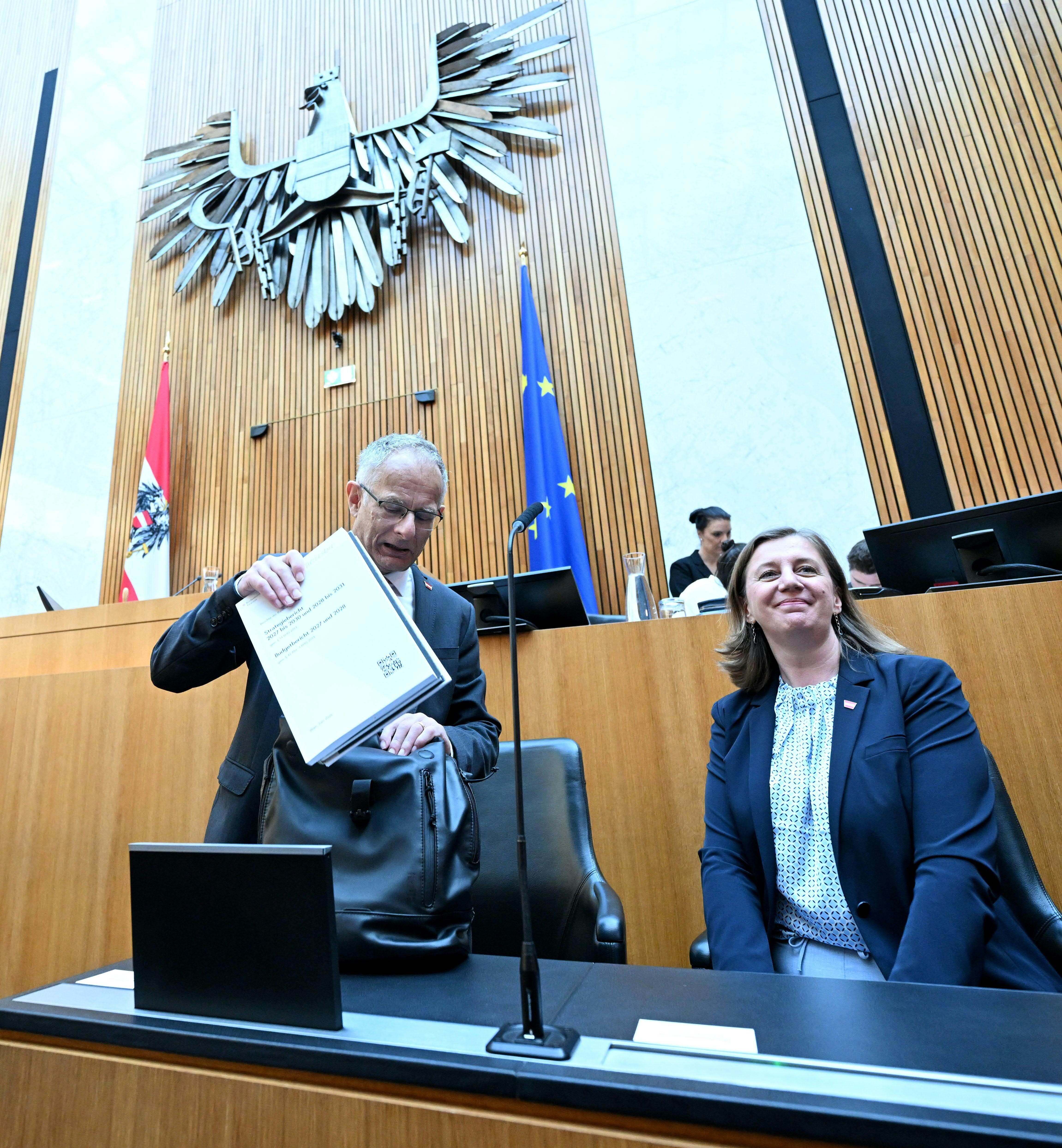 Zwei Personen sitzen im Parlament in Wien vor Nationalrats- und EU-Flagge.