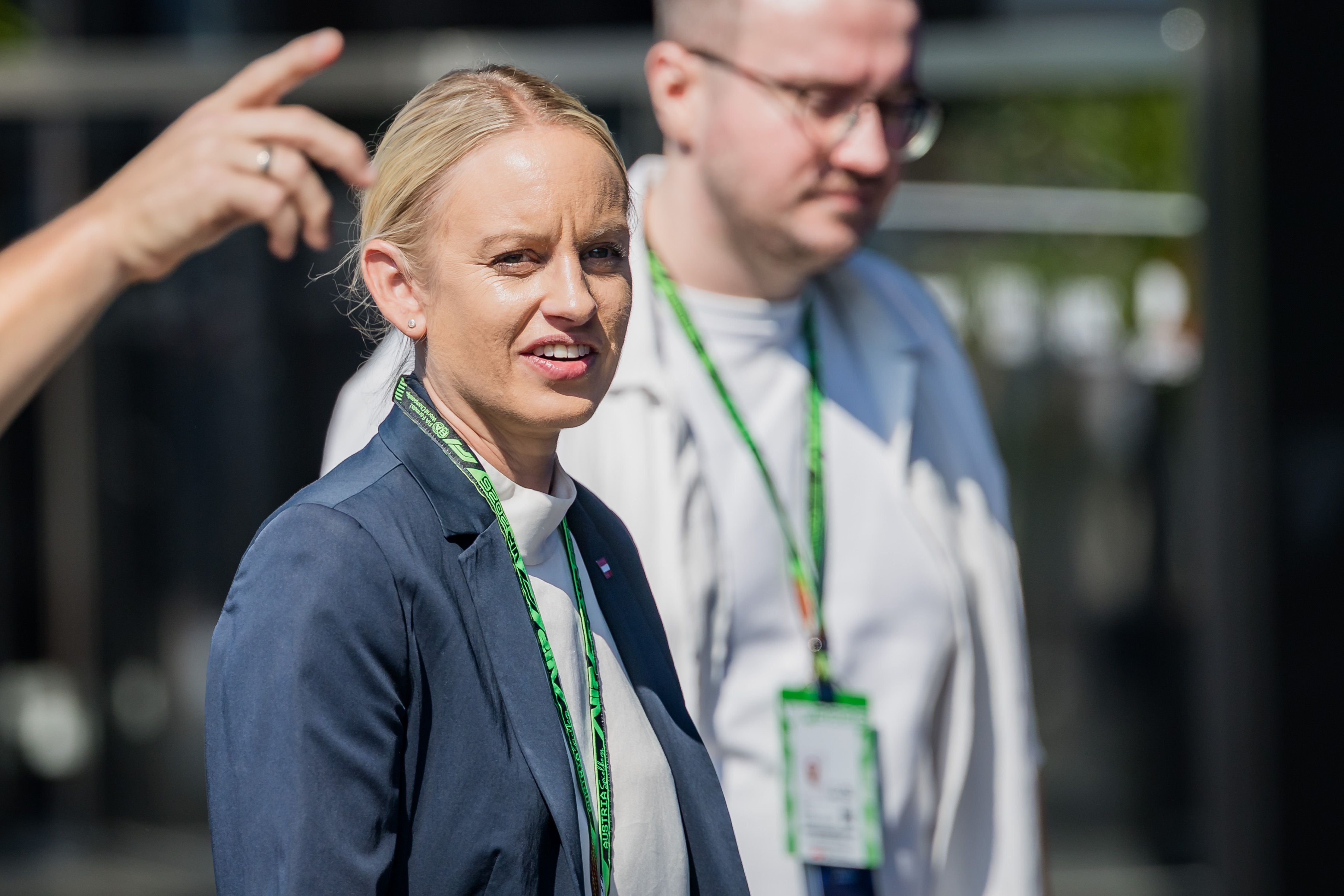 Eine Frau im Anzug mit grünem Ausweisband steht im Freien, im Hintergrund eine zweite Person.